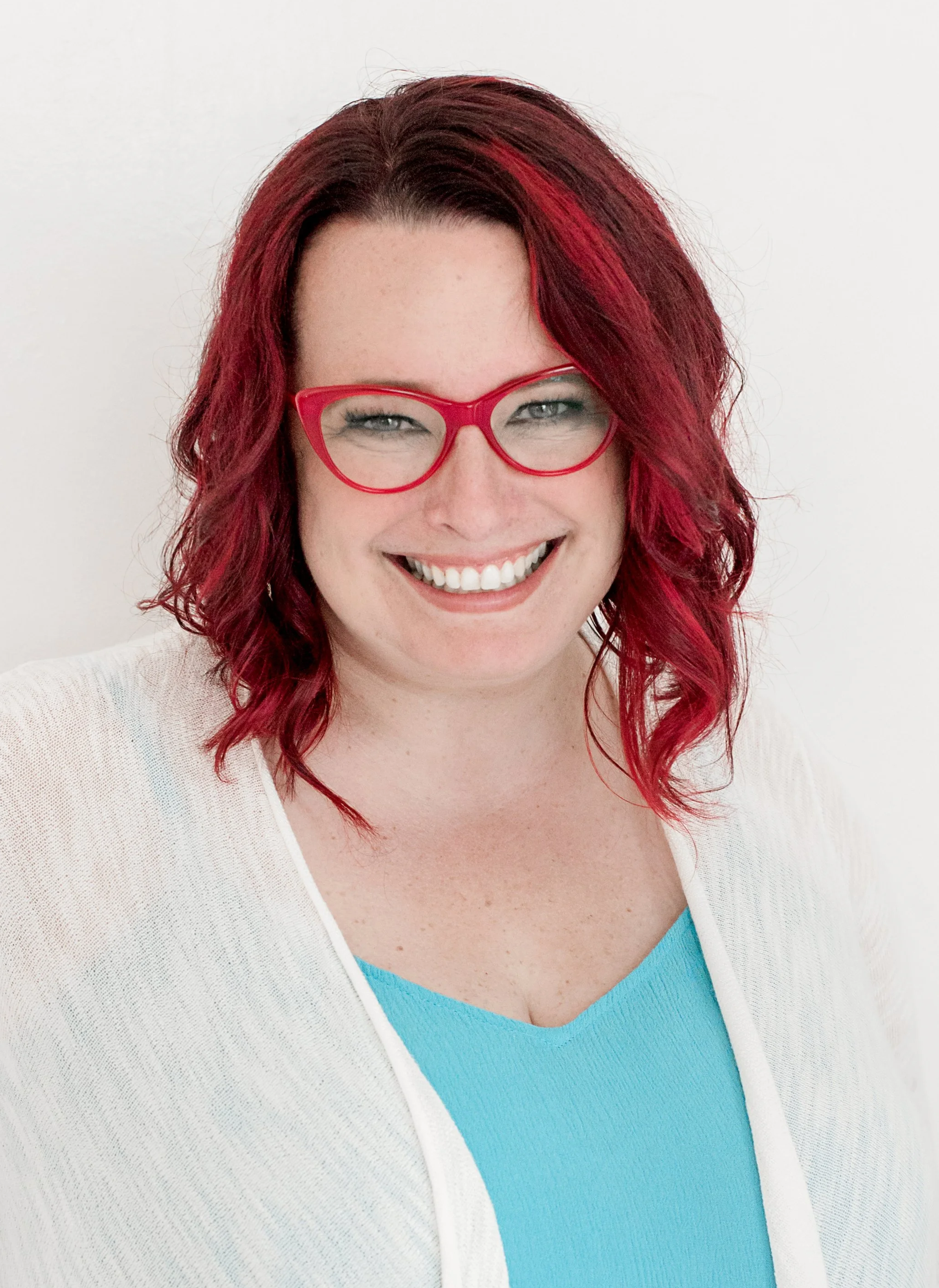 A woman with red hair, red glasses, wearing a white cardigan over a teal top, smiling, against a white background.