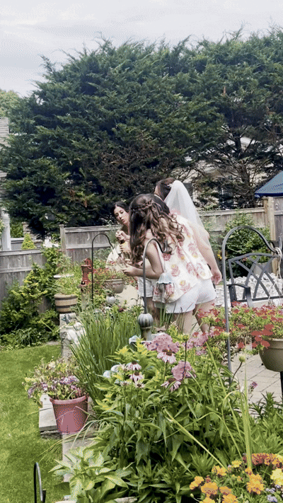 Three women gather in a backyard garden with flowering plants and outdoor furniture, with a large tree and wooden fence in the background.