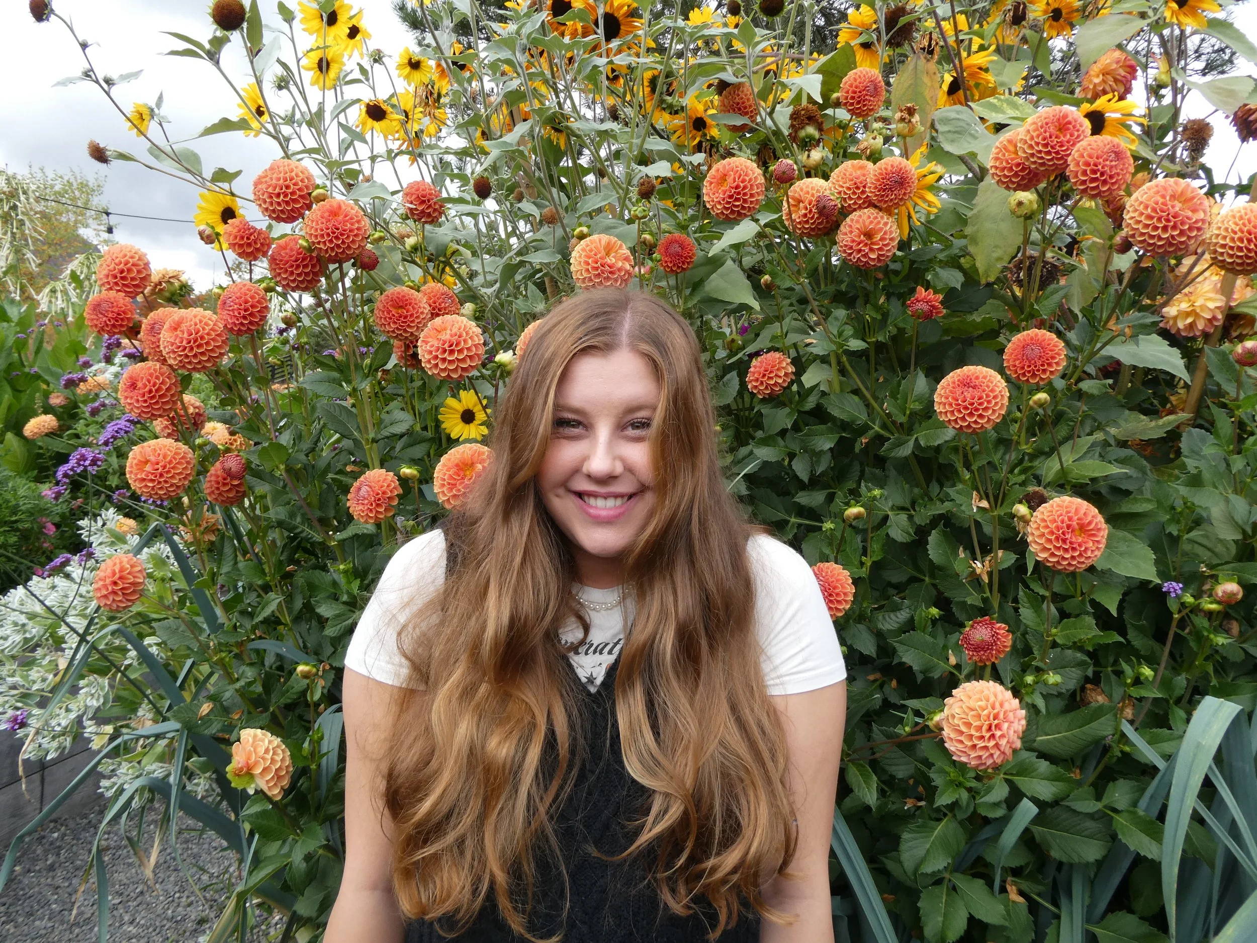 A smiling young woman with long wavy hair stands amidst a colorful flower garden with orange dahlia flowers and yellow sunflowers.