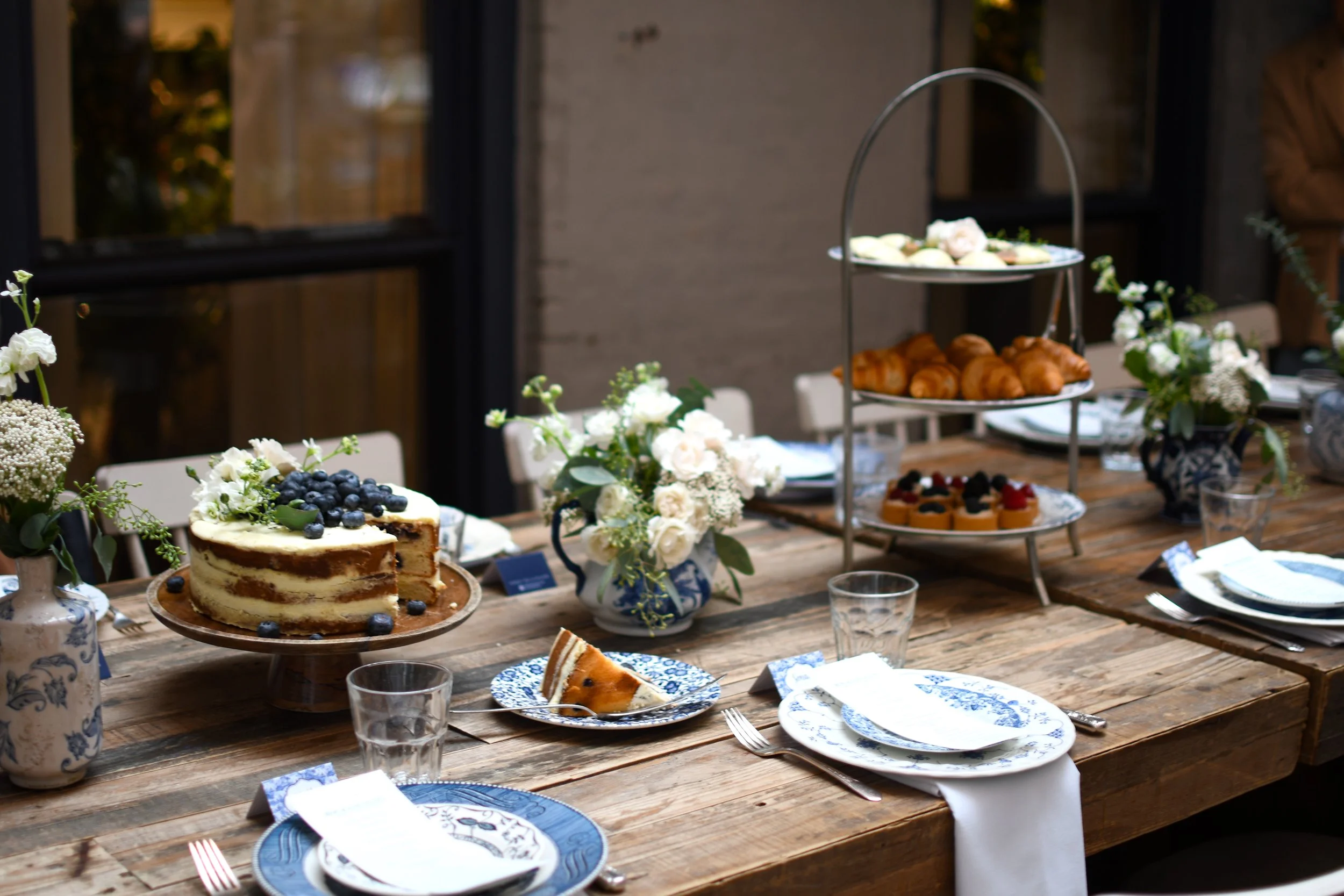 A rustic wooden dining table set with blue and white patterned plates, silverware, empty glasses, and white napkins. The table is decorated with vases of white flowers. There is a three-tiered serving stand with croissants, pastries, and small tarts, and a blue-iced layered cake topped with blueberries and white flowers. A slice of cake is on a small plate.