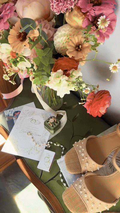 A top view of a floral bouquet with pink, white, and beige flowers, jewelry including a pearl necklace, a card, a small ring box, a pair of tan high-heeled shoes with pearl embellishments, and a photo on a green cushioned table.