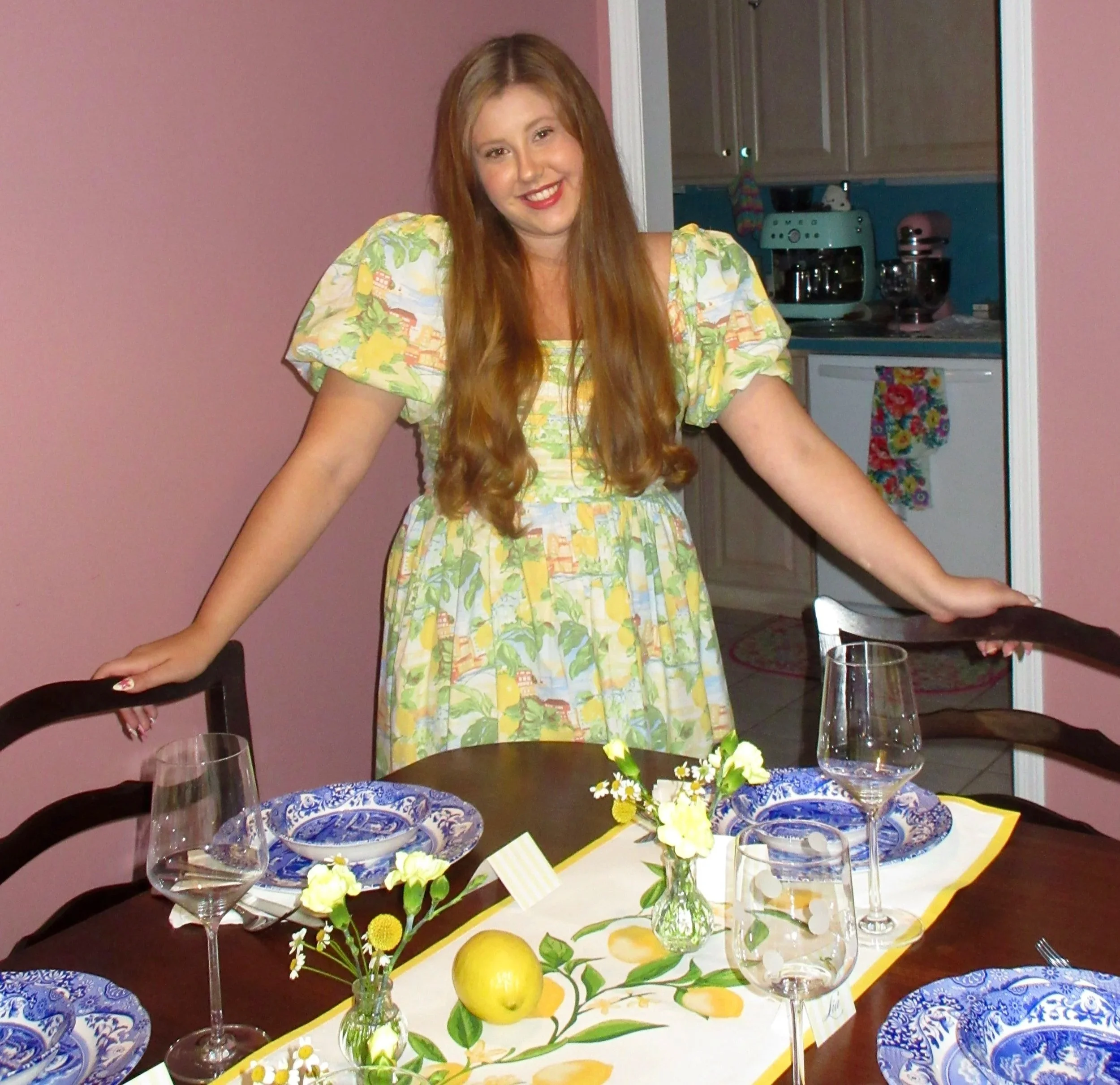 A young woman with long hair smiling at a table set for a dinner party, with floral arrangements and lemons, in a dining room with pink walls and visible kitchen in the background.