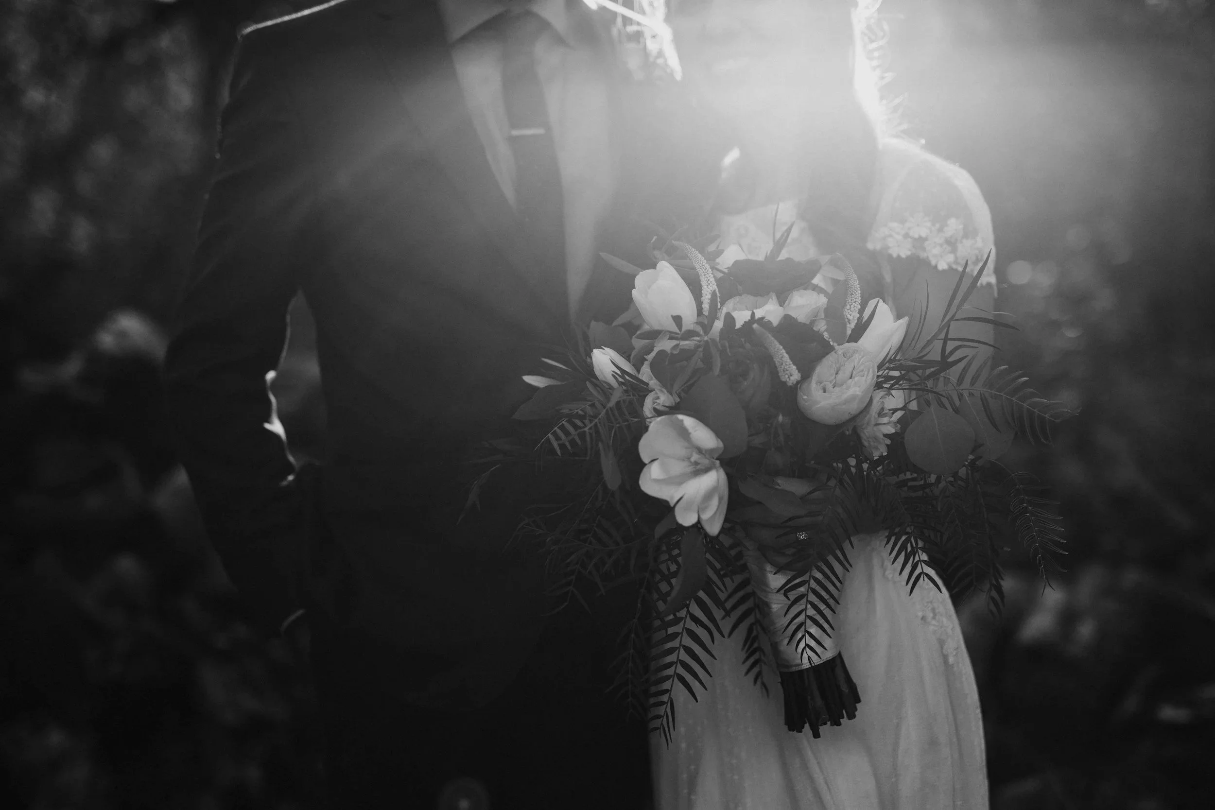 A groom in a tuxedo holding a bouquet of flowers depicted on a black-and-white photo, with sunlight shining behind.