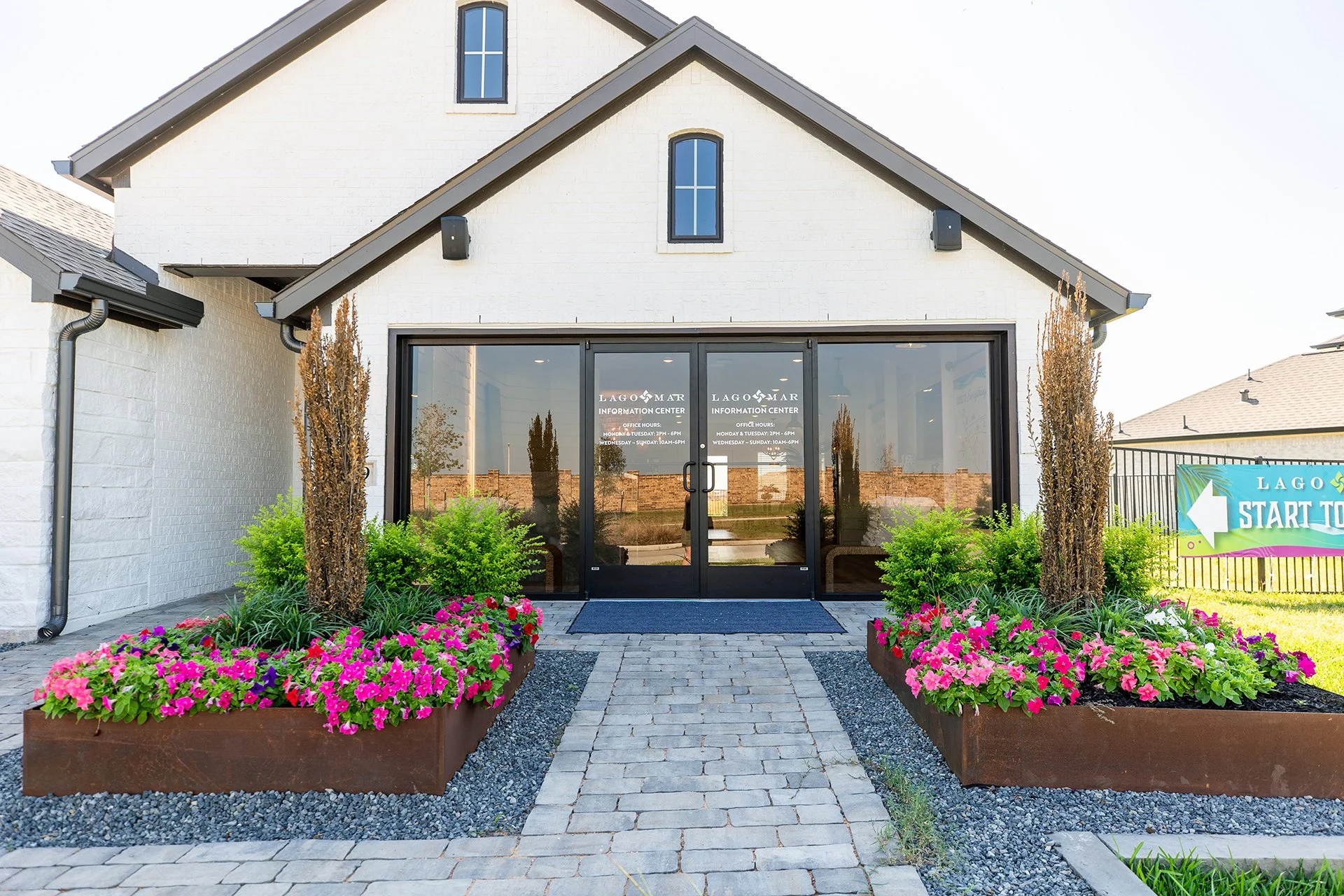 Front entrance of a white brick building with glass doors, potted plants, flowering bushes, and paved walkway.