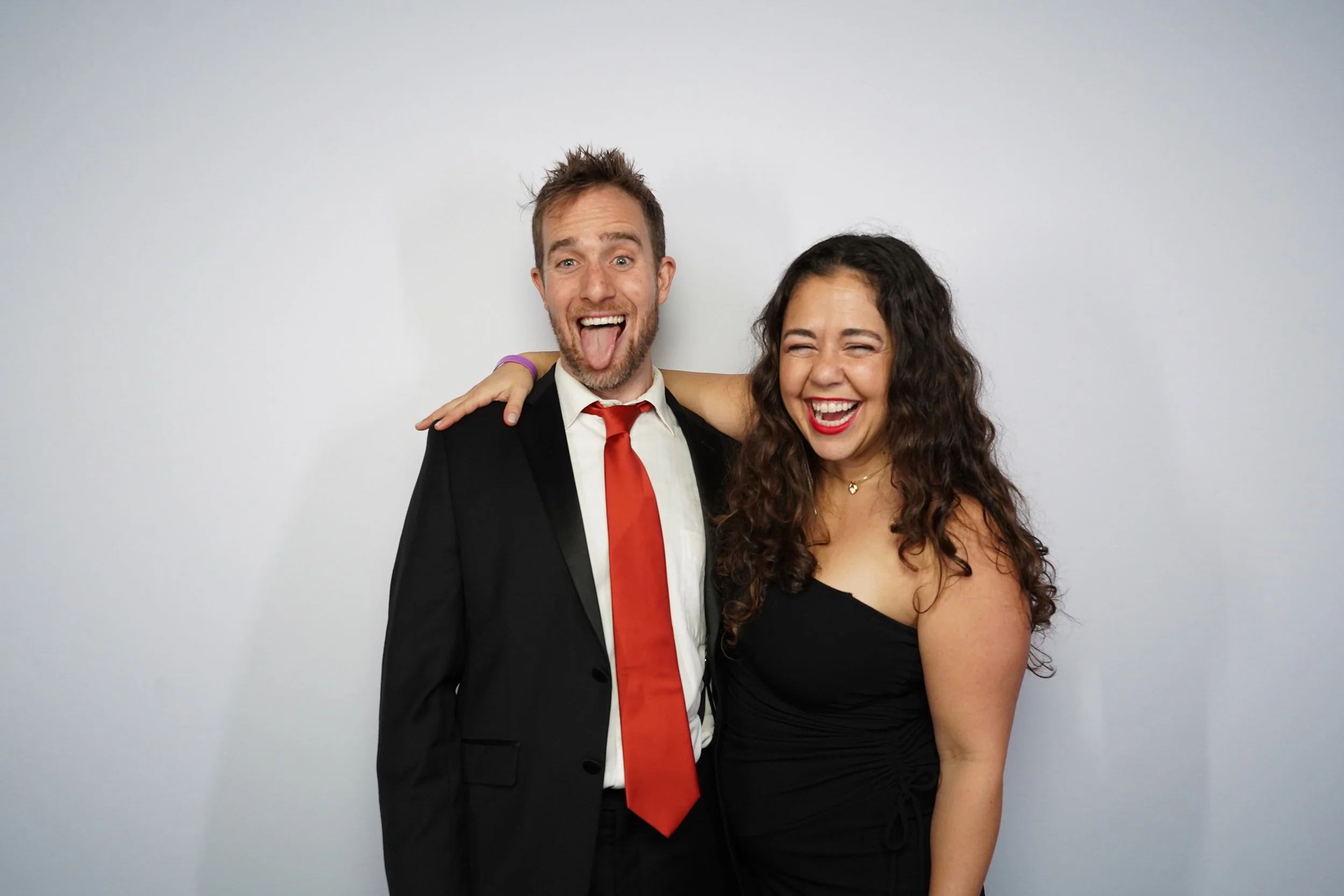 A man in a black suit and red tie with a beard, and a woman in a black dress with curly hair, standing together and laughing in front of a plain white background.