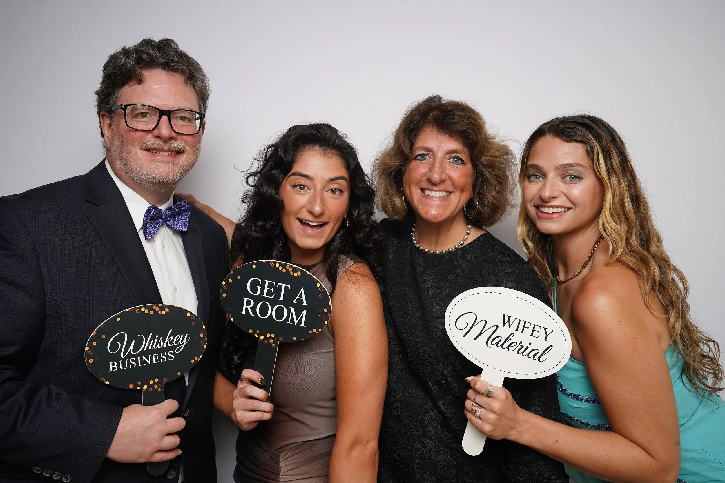 Group of four people dressed formally, holding humorous signs that read 'Whiskey Business,' 'Get a Room,' and 'Wifey Material,' smiling against a plain white background.