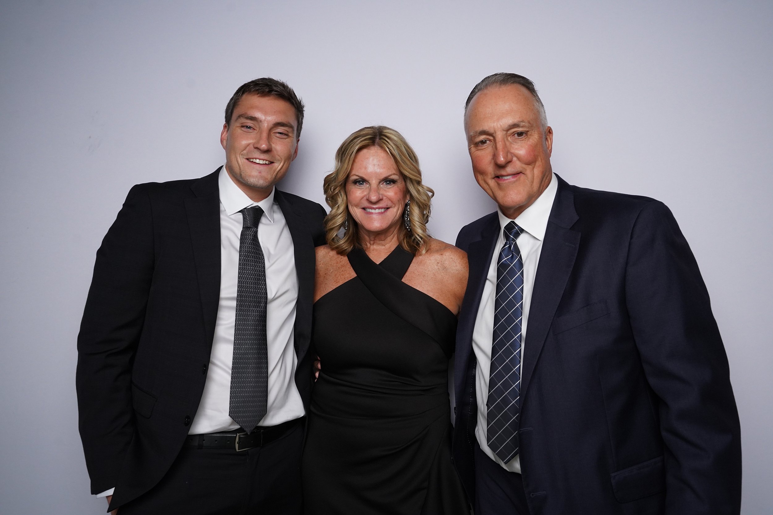 Three people in formal attire standing close together and smiling at the camera against a plain light gray background.