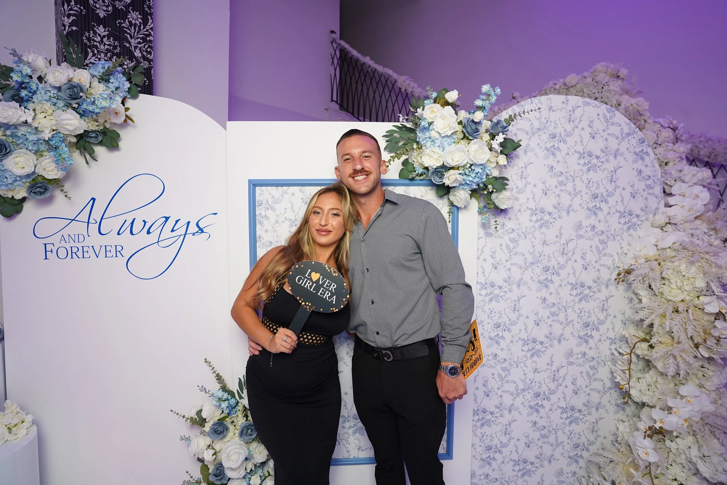 A couple posing at a wedding celebration. The woman is holding a sign that says 'I love ever girl era' and they are standing in front of a decorated backdrop with flowers and the words 'Always and Forever'.