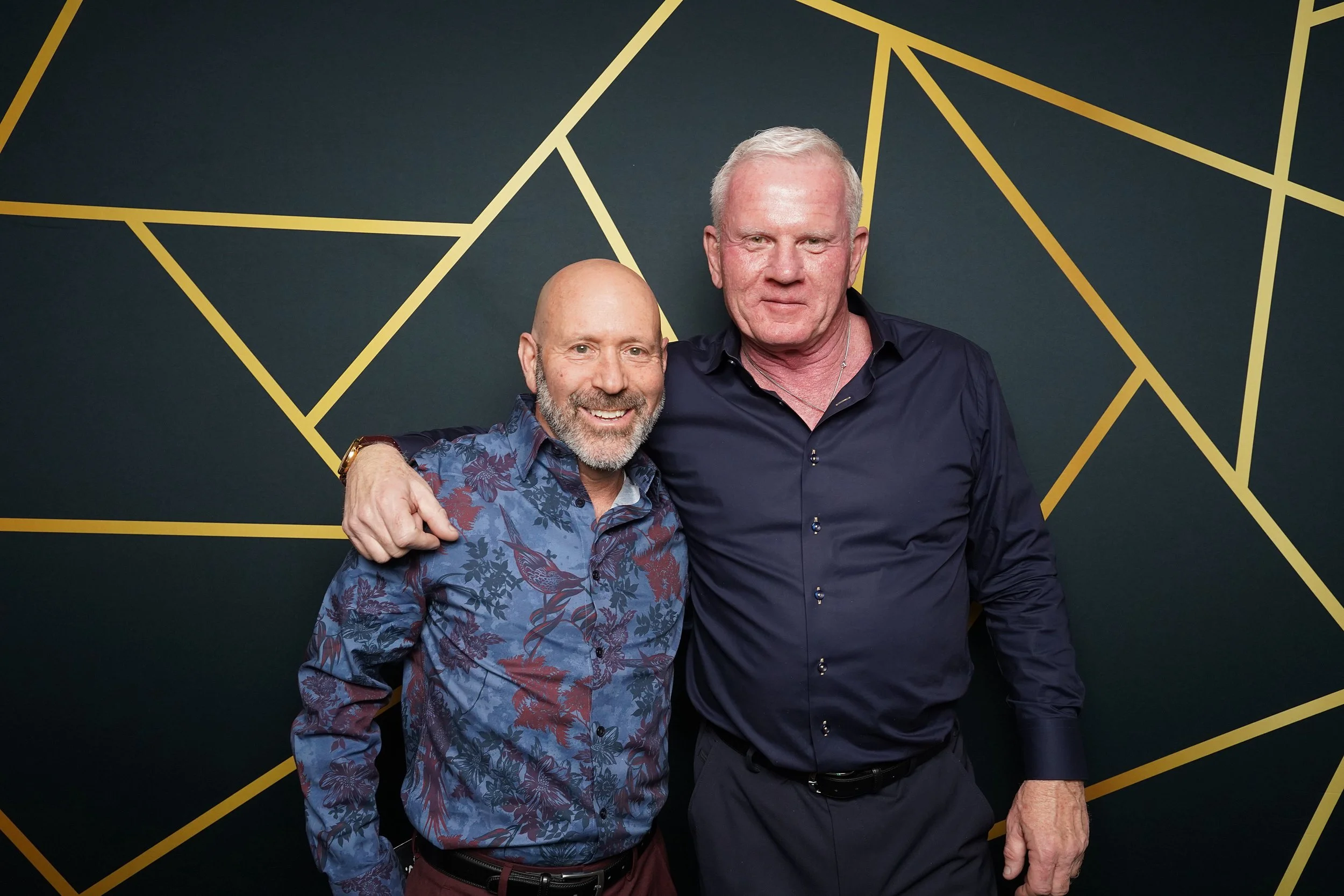 Two men standing together with their arms around each other's shoulders, smiling at the camera, against a dark wall with gold geometric lines.