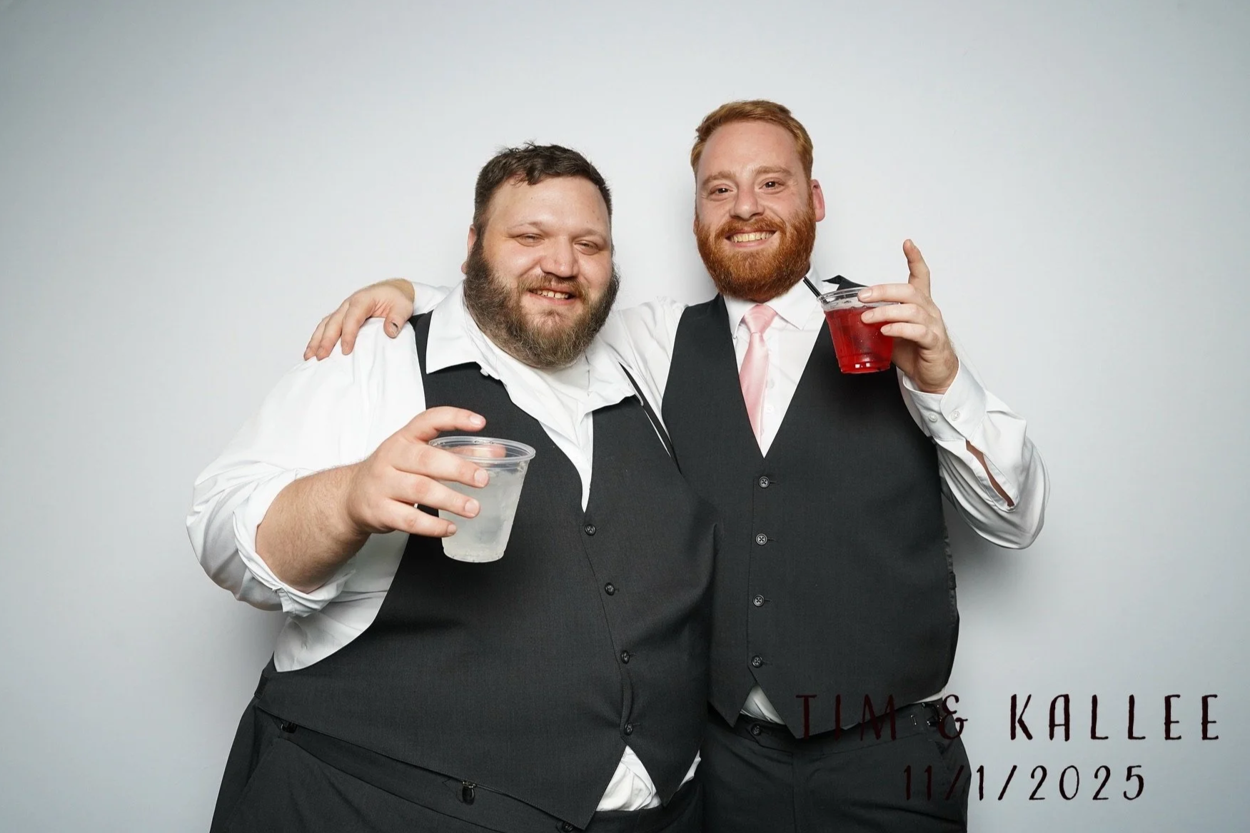 Two men in formal attire, with one having a beard and the other having reddish hair and a beard, standing close together with their arms around each other, smiling, holding drinks, in front of a plain background, with text in the bottom right corner 