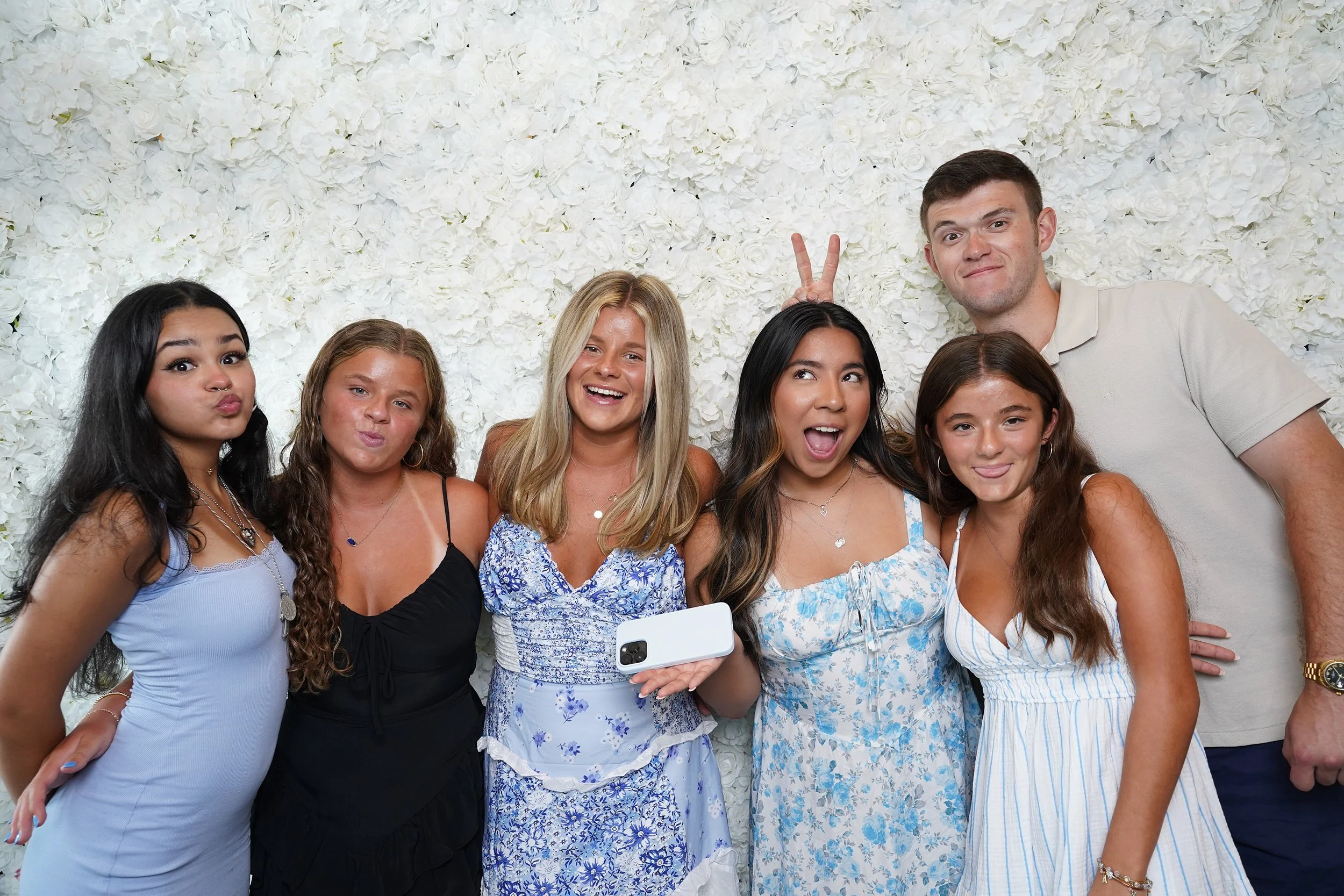 Group of six young people celebrating, posing playfully in front of a white floral backdrop, with one person making bunny ears behind another's head.
