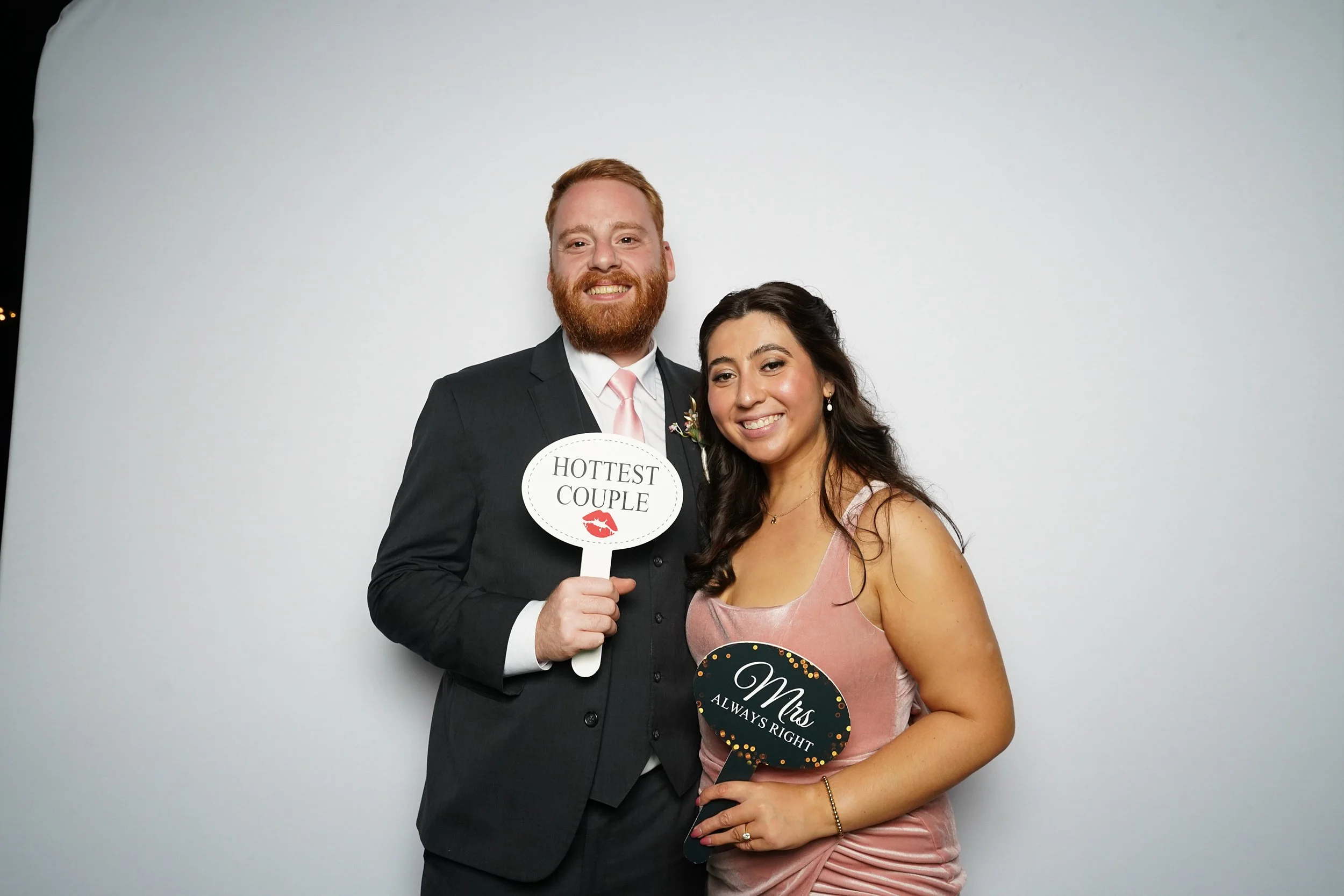 A smiling couple dressed in formal attire at an event, holding signs that read 'Hottest Couple' and 'Ms Always Right' in front of a plain background.