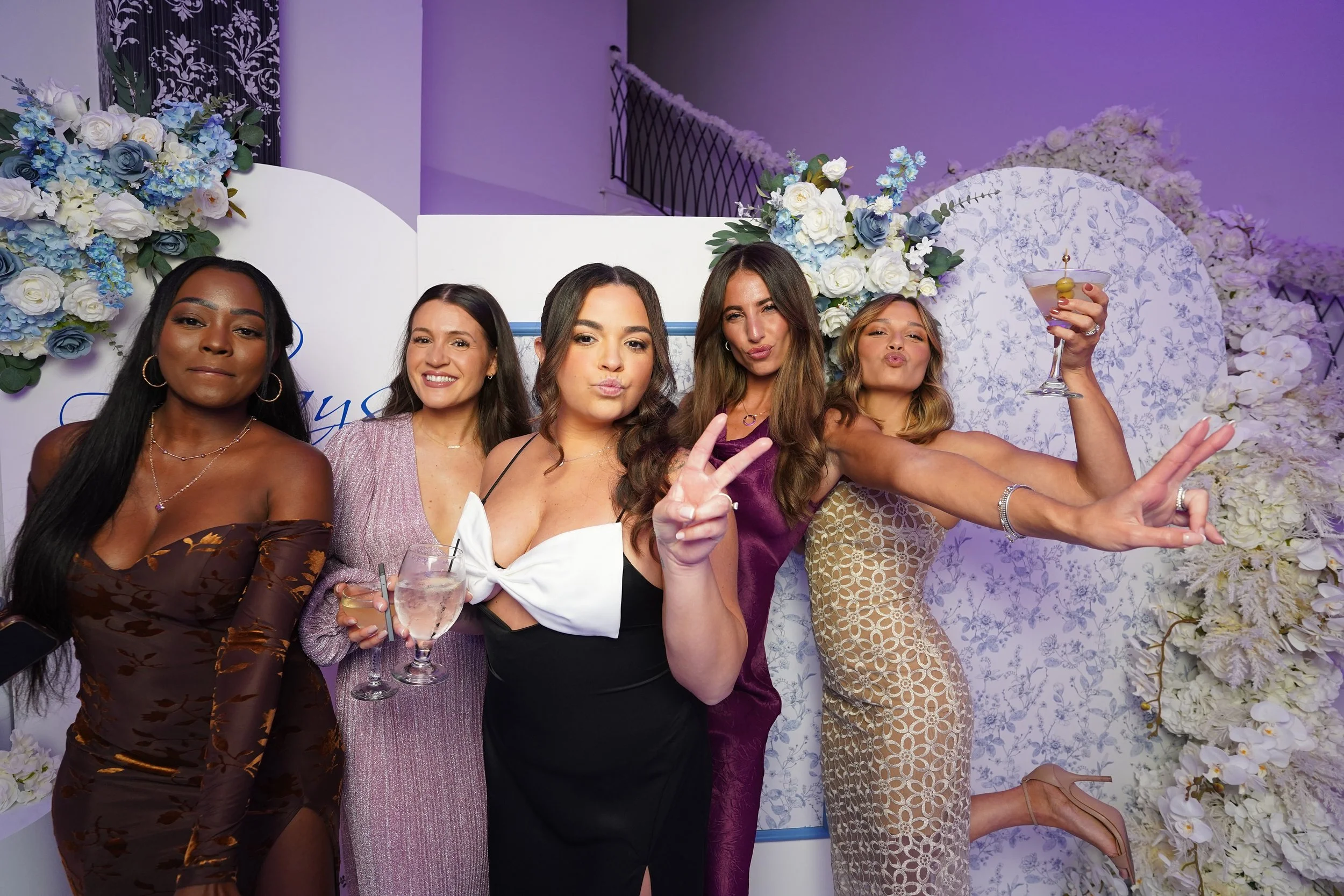 Five women at a celebration, posing with drinks, in front of a floral backdrop with white and blue flowers and a white screen.