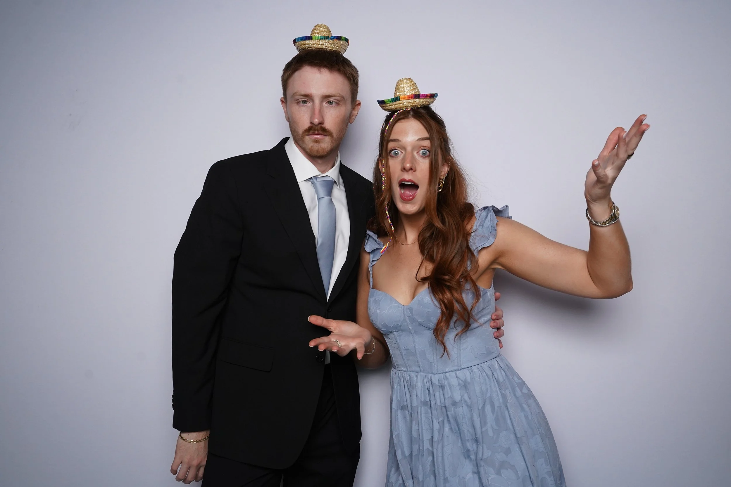 A man and woman standing side by side against a plain light background, both wearing small colorful sombreros on their heads. The man is dressed in a black suit and light blue tie, with a serious expression. The woman is wearing a light blue dress, h