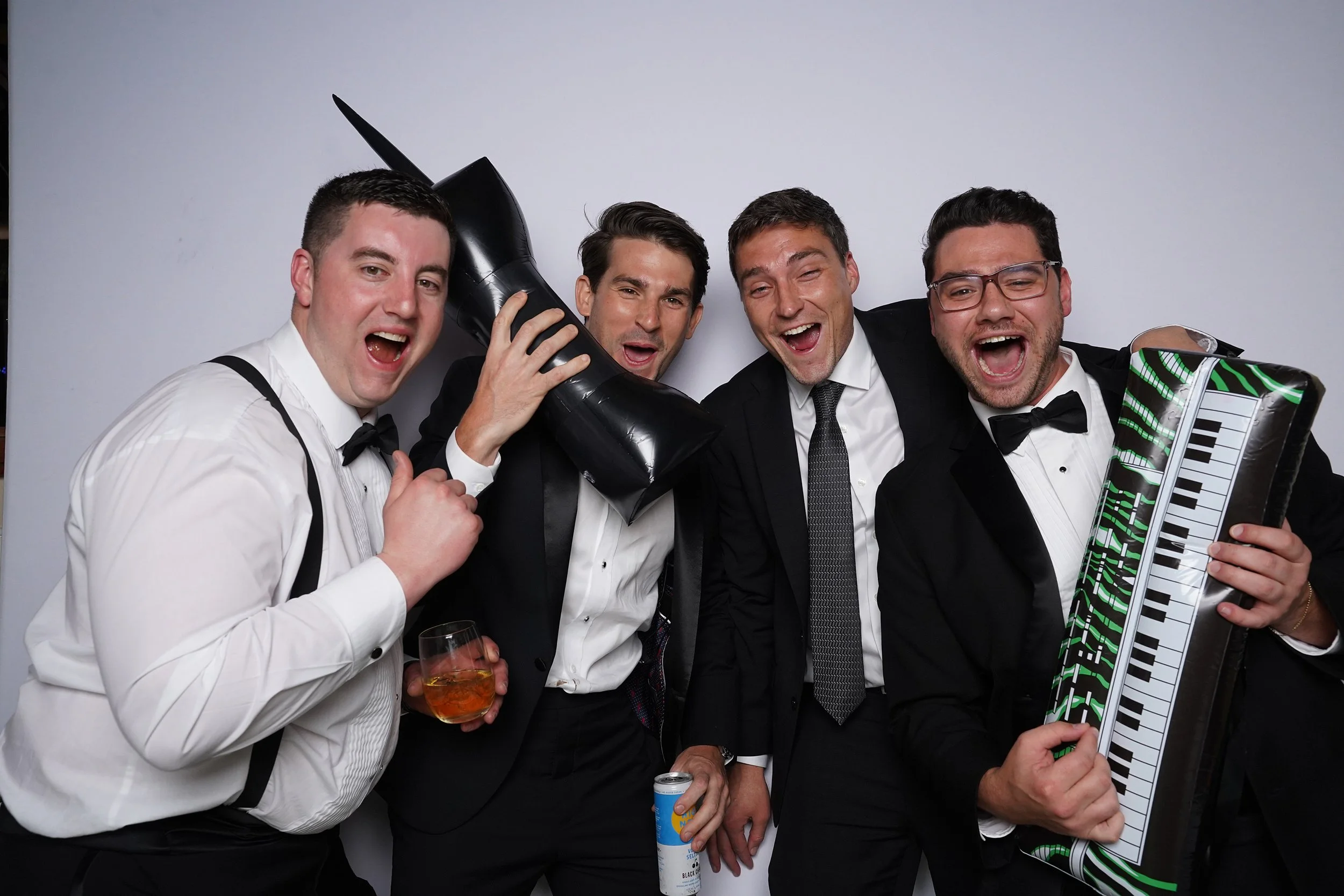 Four men in tuxedos celebrating at a party, holding a toy ak-47, a large inflatable shoe, a drink, and a keyboard-shaped box.