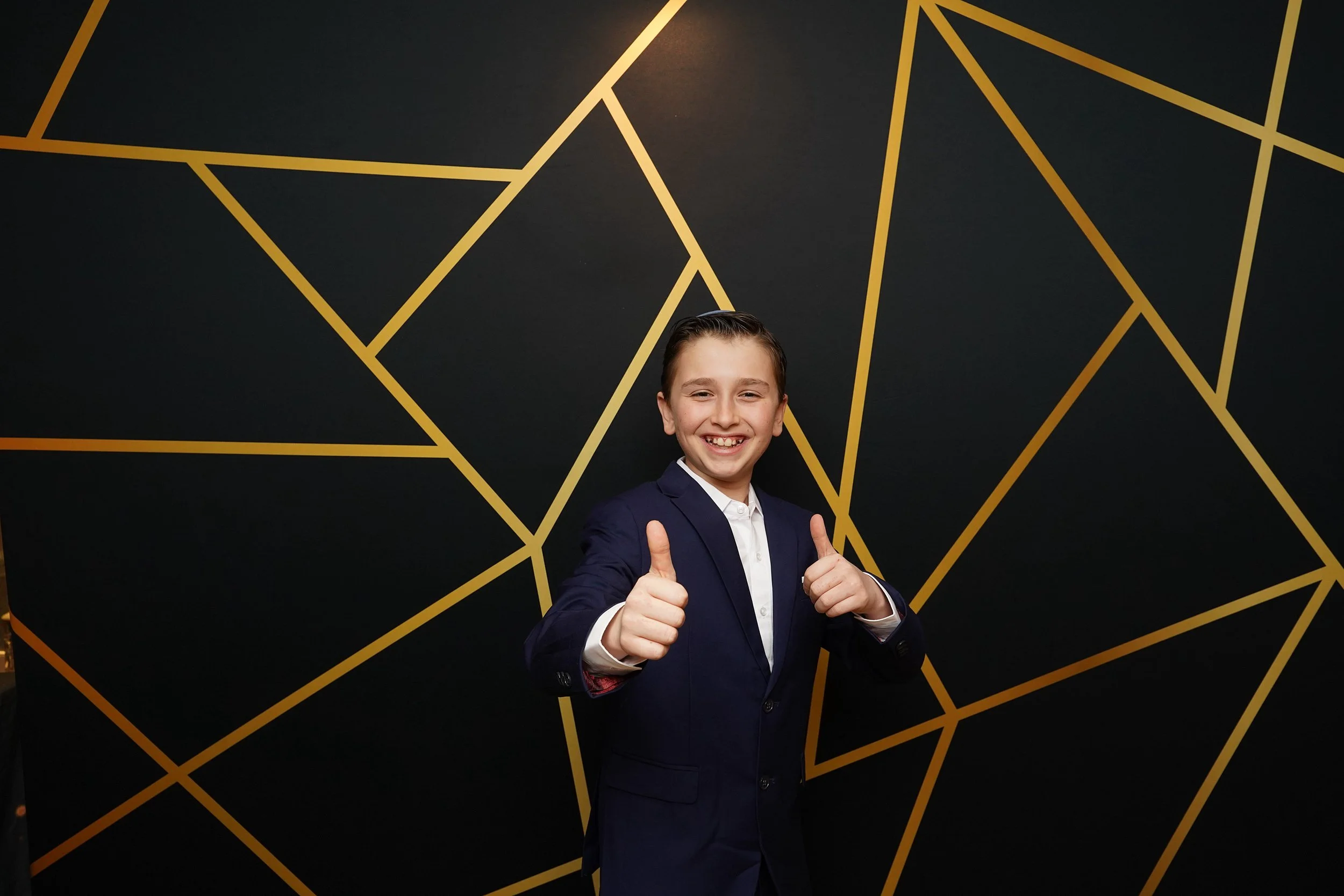 A young boy in a navy suit smiling and giving a thumbs-up in front of a black wall with gold geometric lines.