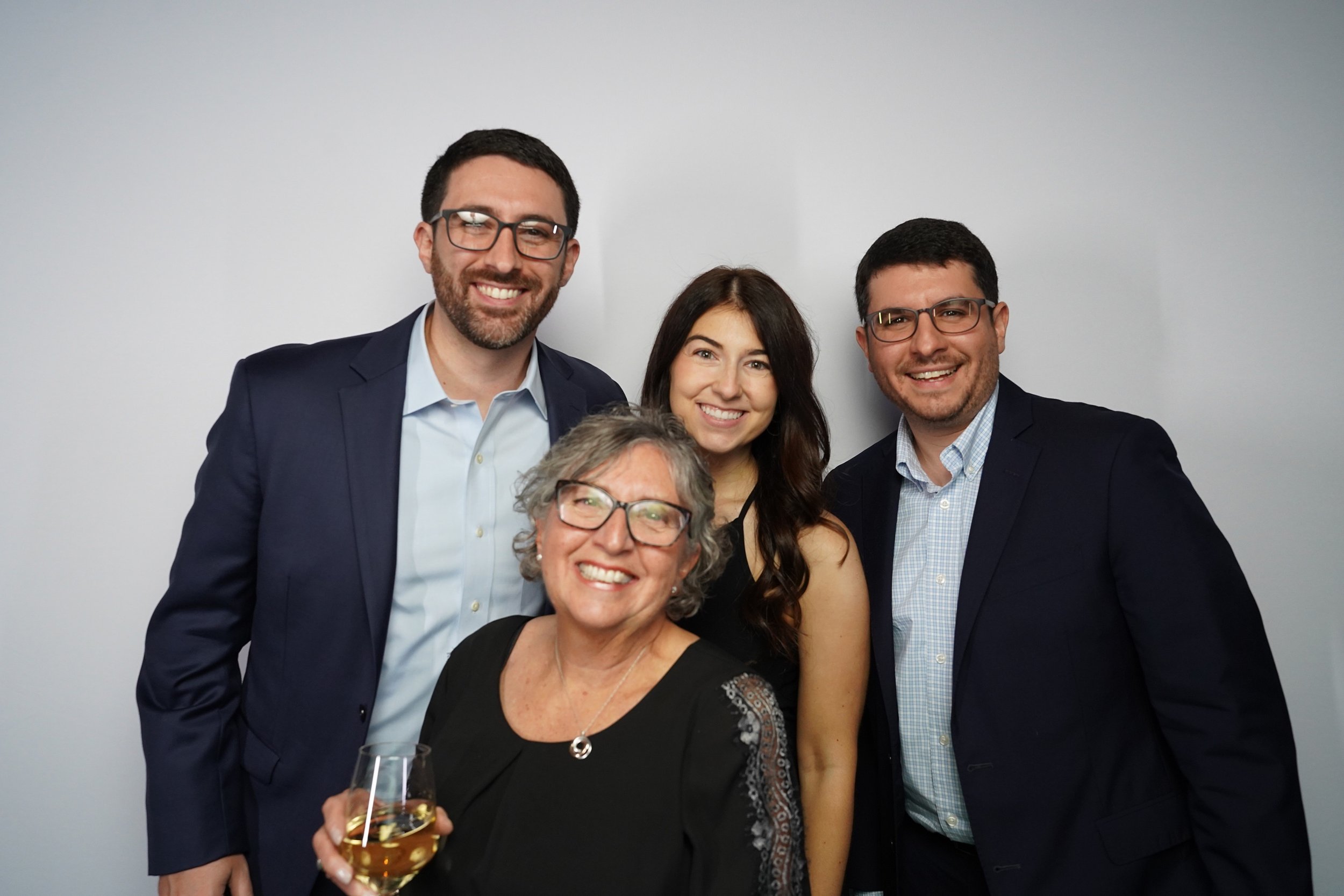 Group of four adults and one older woman smiling and posing together at a social event, with the older woman holding a glass of white wine, against a plain white wall.