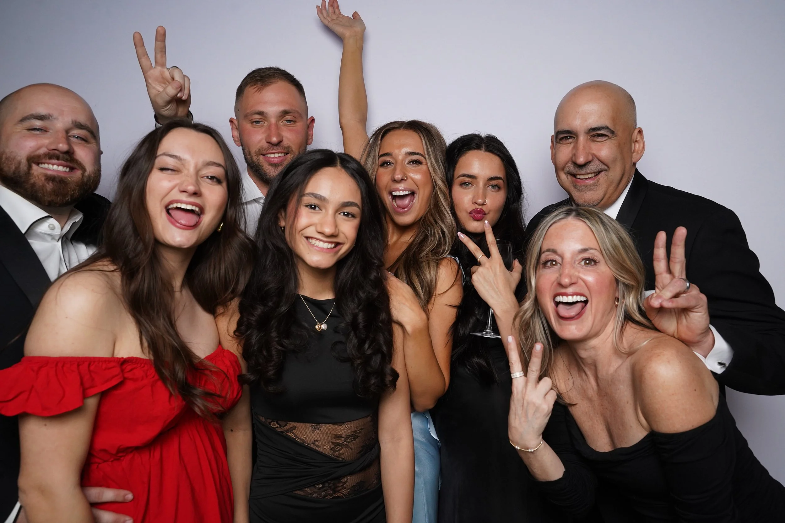 Group of nine people celebrating and posing for a photo, smiling, making peace signs, and showing playful gestures against a plain background.