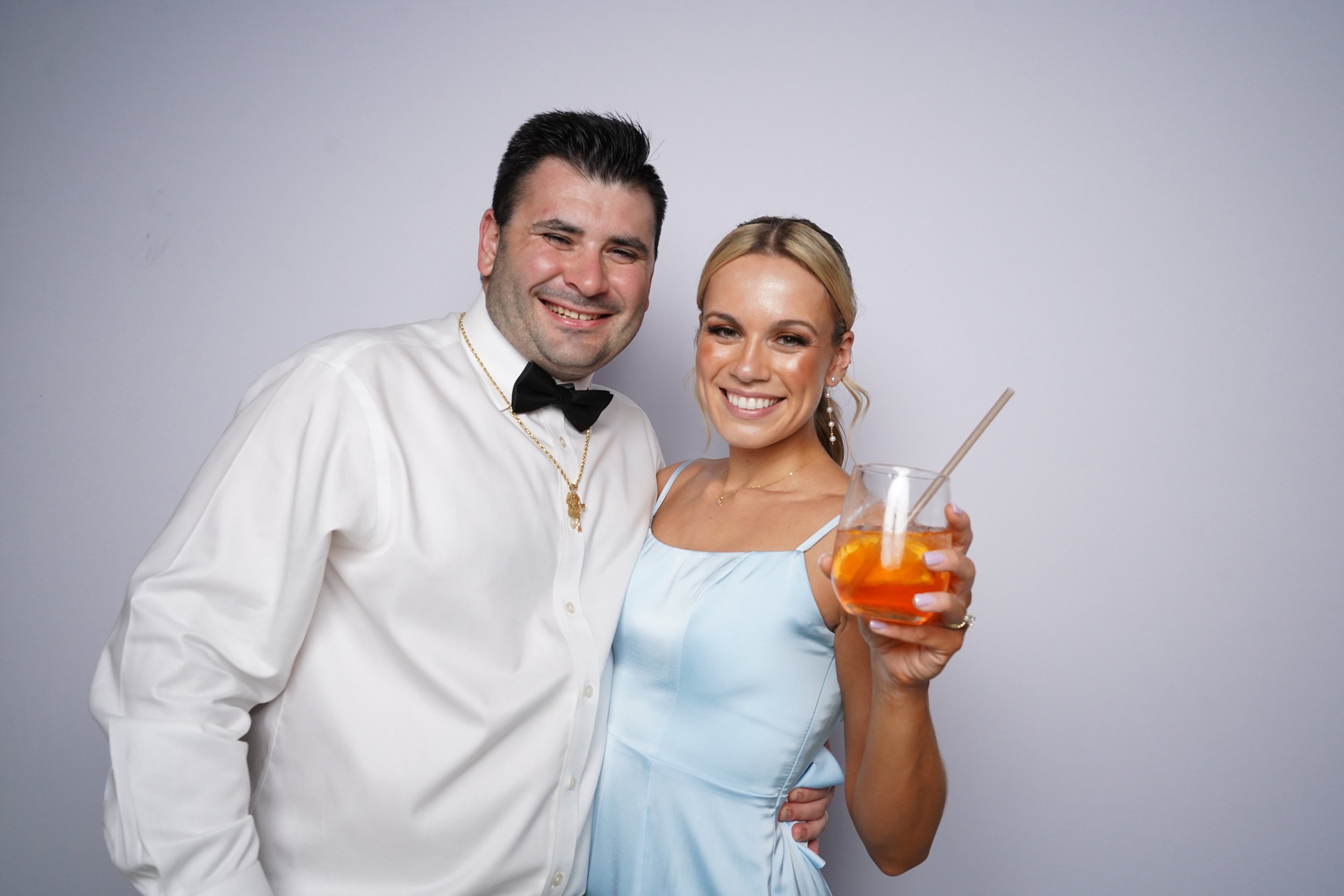 A smiling man dressed in a white shirt with a black bow tie and a woman in a light blue dress holding a glass of cocktail, both standing close together in front of a plain gray background.