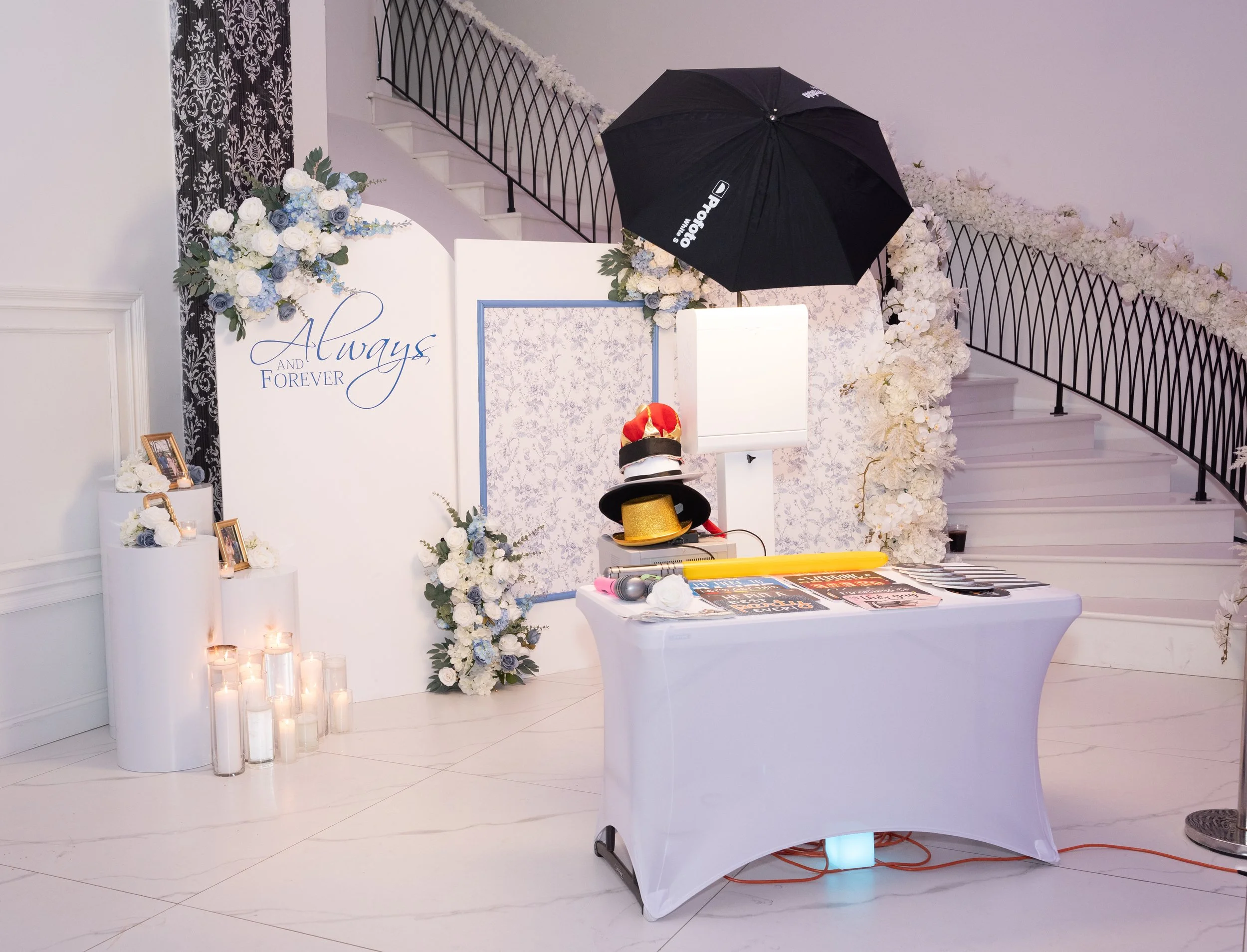 Decorative wedding photo booth area with white and light blue floral arrangements, candles, photo frames, a black umbrella, and hats on a table. The backdrop reads "Always and Forever." A staircase with black railing is visible in the background.