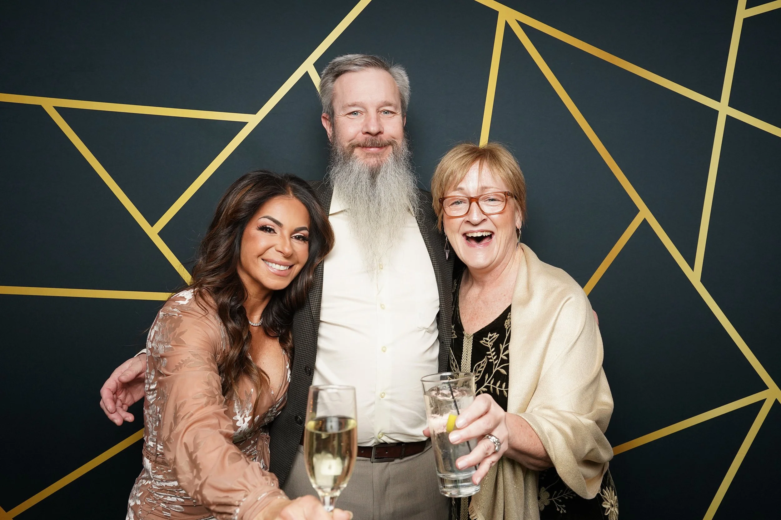 Three people smiling and celebrating at an event, holding drinks, standing in front of a dark background with gold geometric lines.