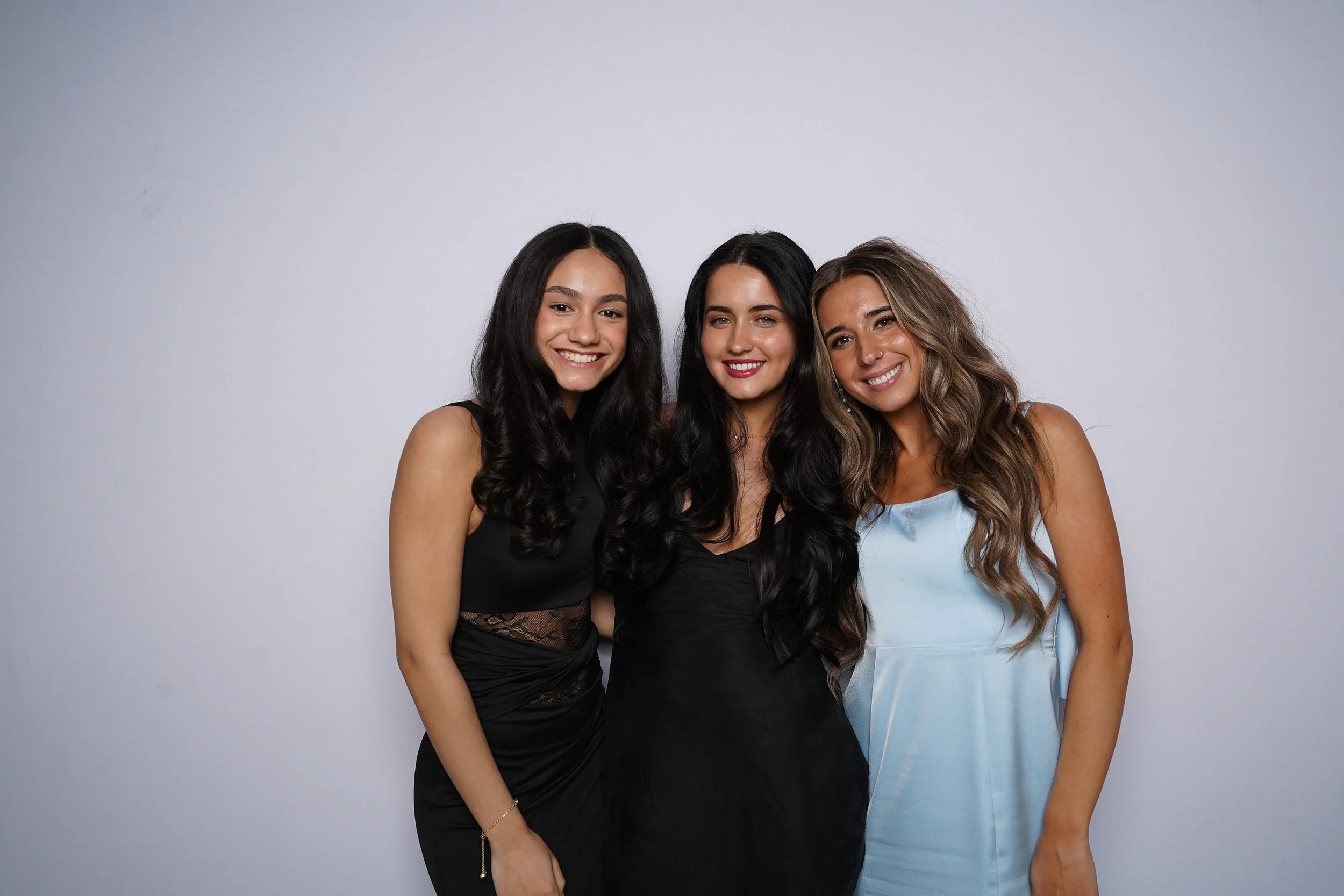 Three young women standing close together, smiling, against a plain white background. They are dressed in black, black, and light blue dresses.