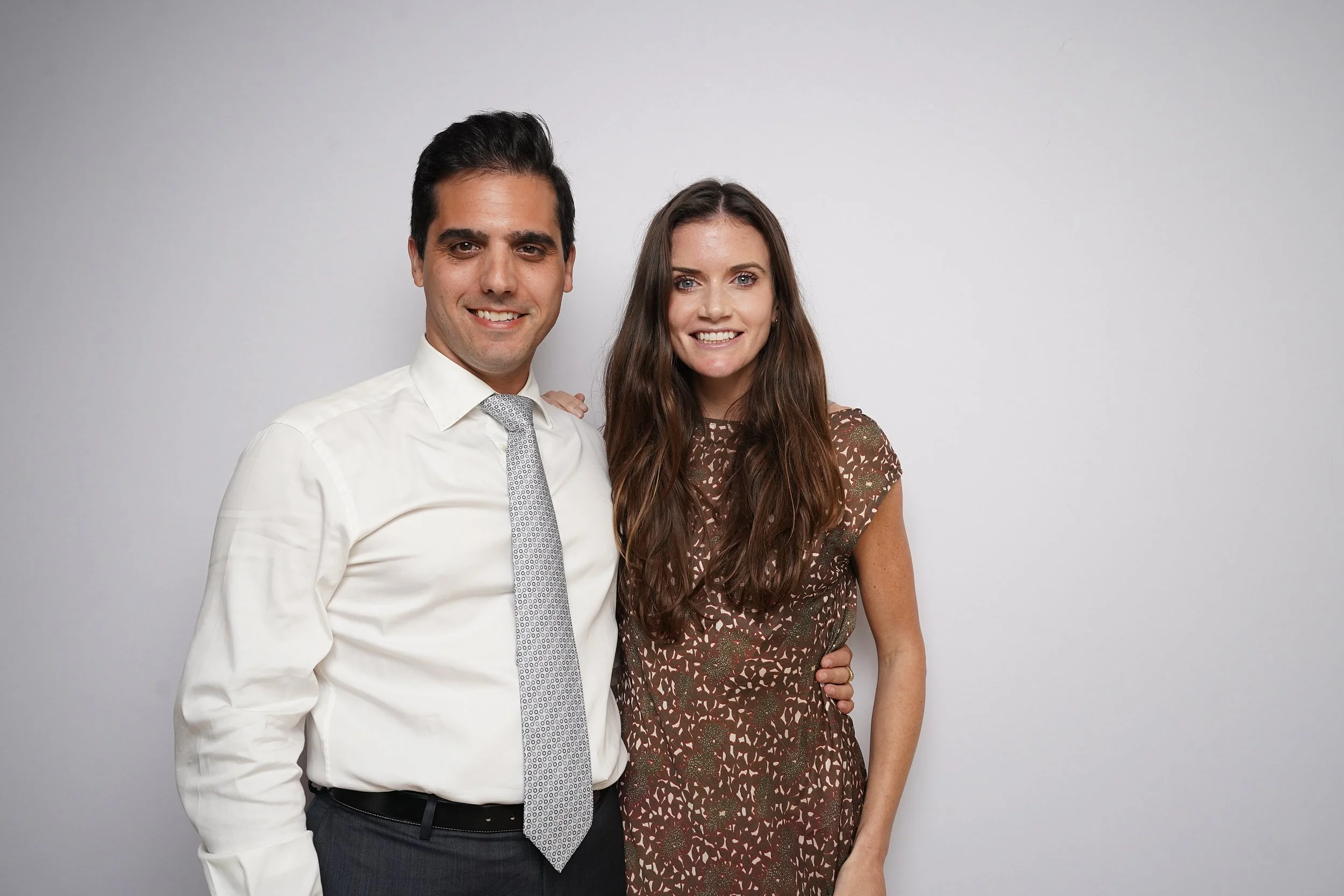 A man in a white shirt and tie standing next to a woman in a brown patterned dress, both smiling at the camera against a plain white background.