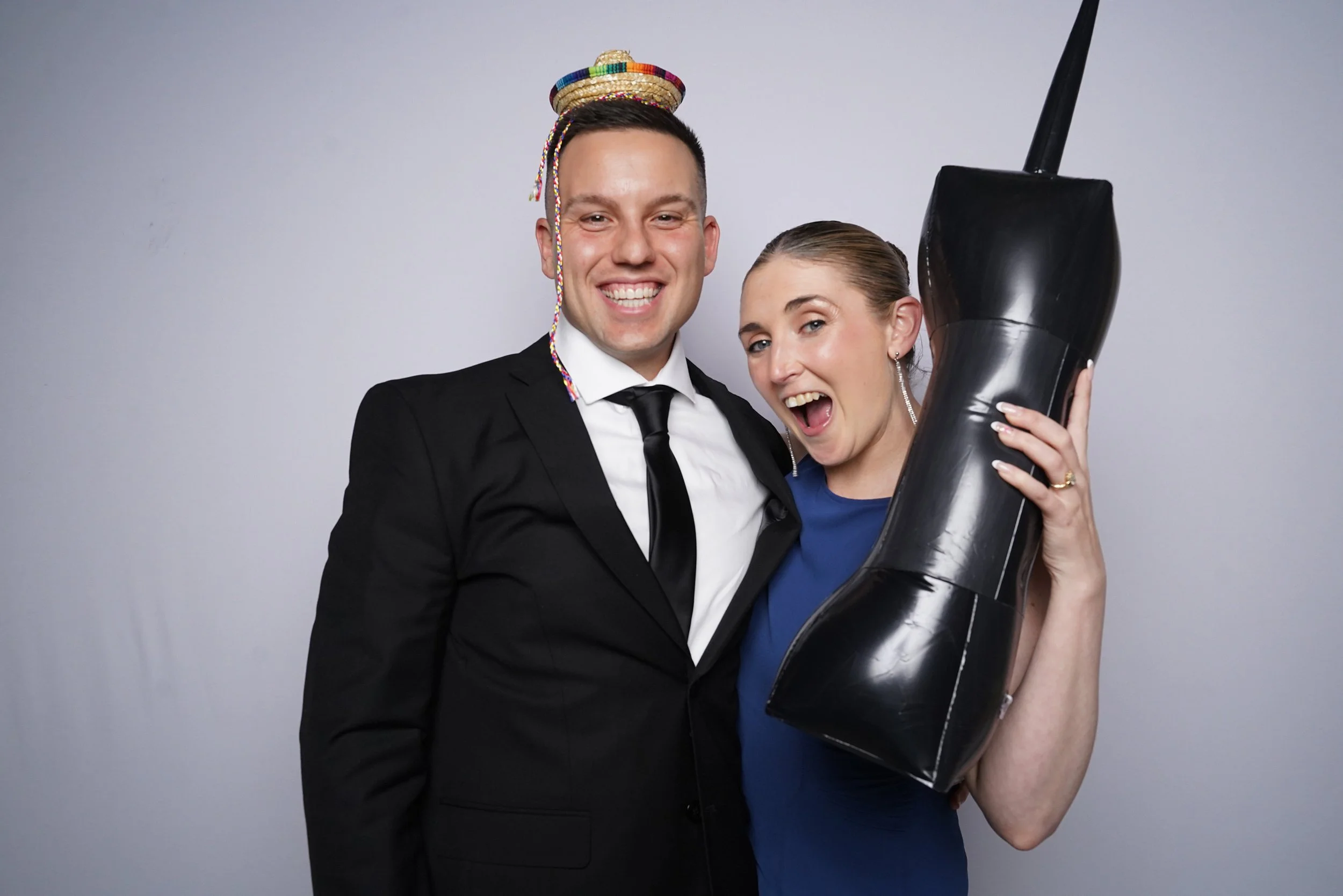 A man and woman celebrating together, with the man wearing a party hat on his head and a black suit, and the woman holding a large black party horn, both smiling happily.