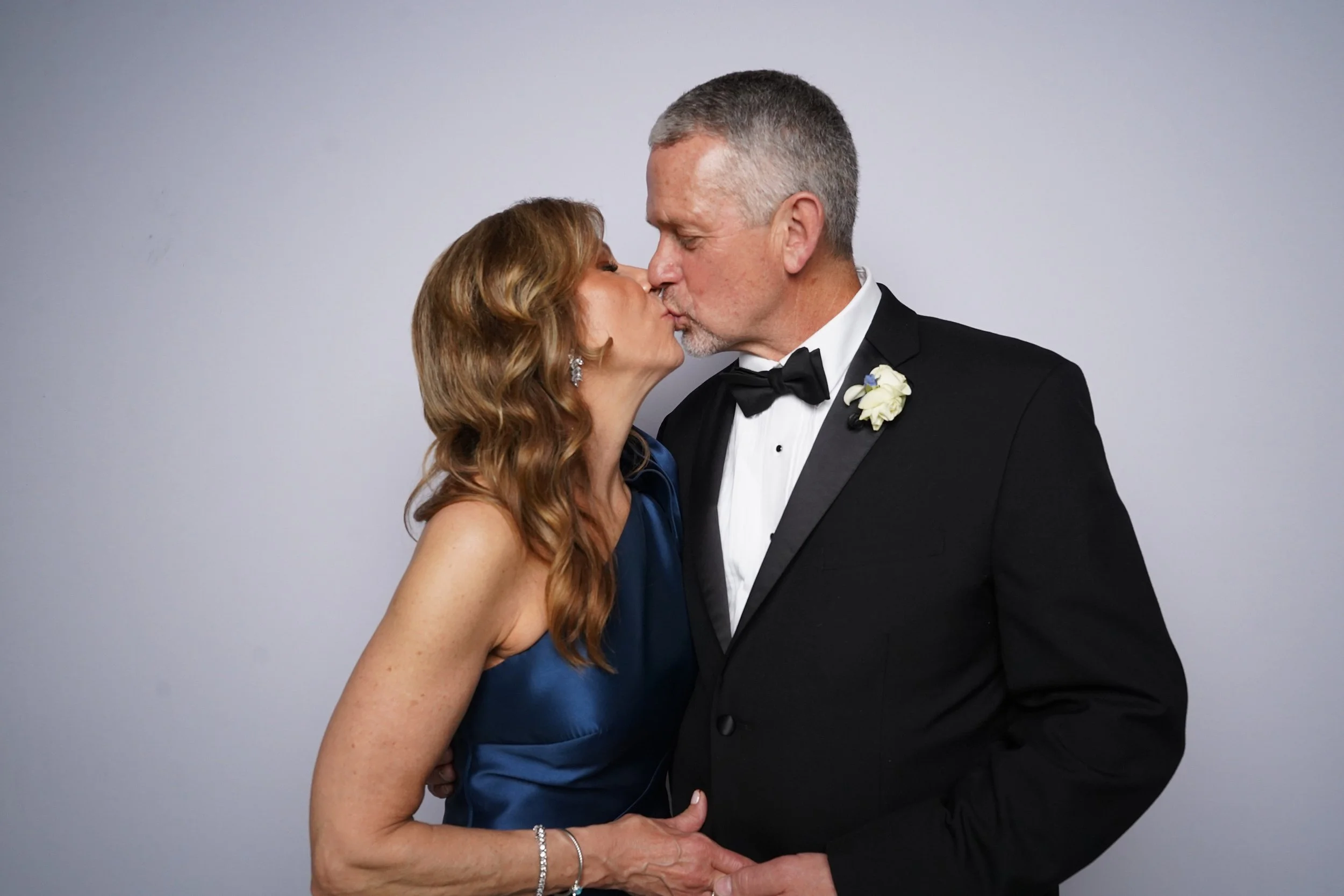 A middle-aged couple in formal attire sharing a kiss, with the woman in a navy blue dress and the man in a black tuxedo, against a plain background.
