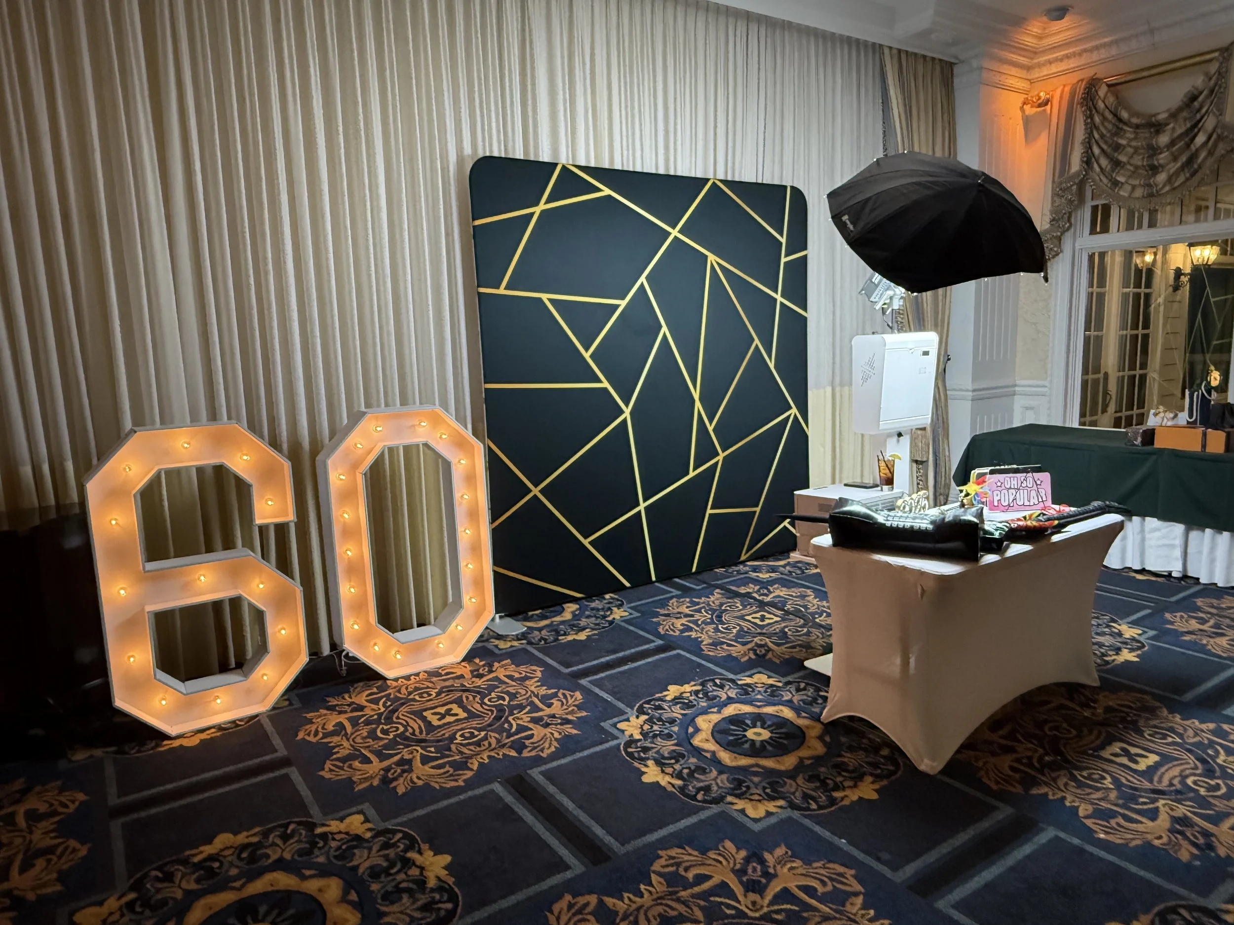 Decorative photo booth setup with illuminated '50' sign, black and gold geometric backdrop, and photography equipment in an elegant room.