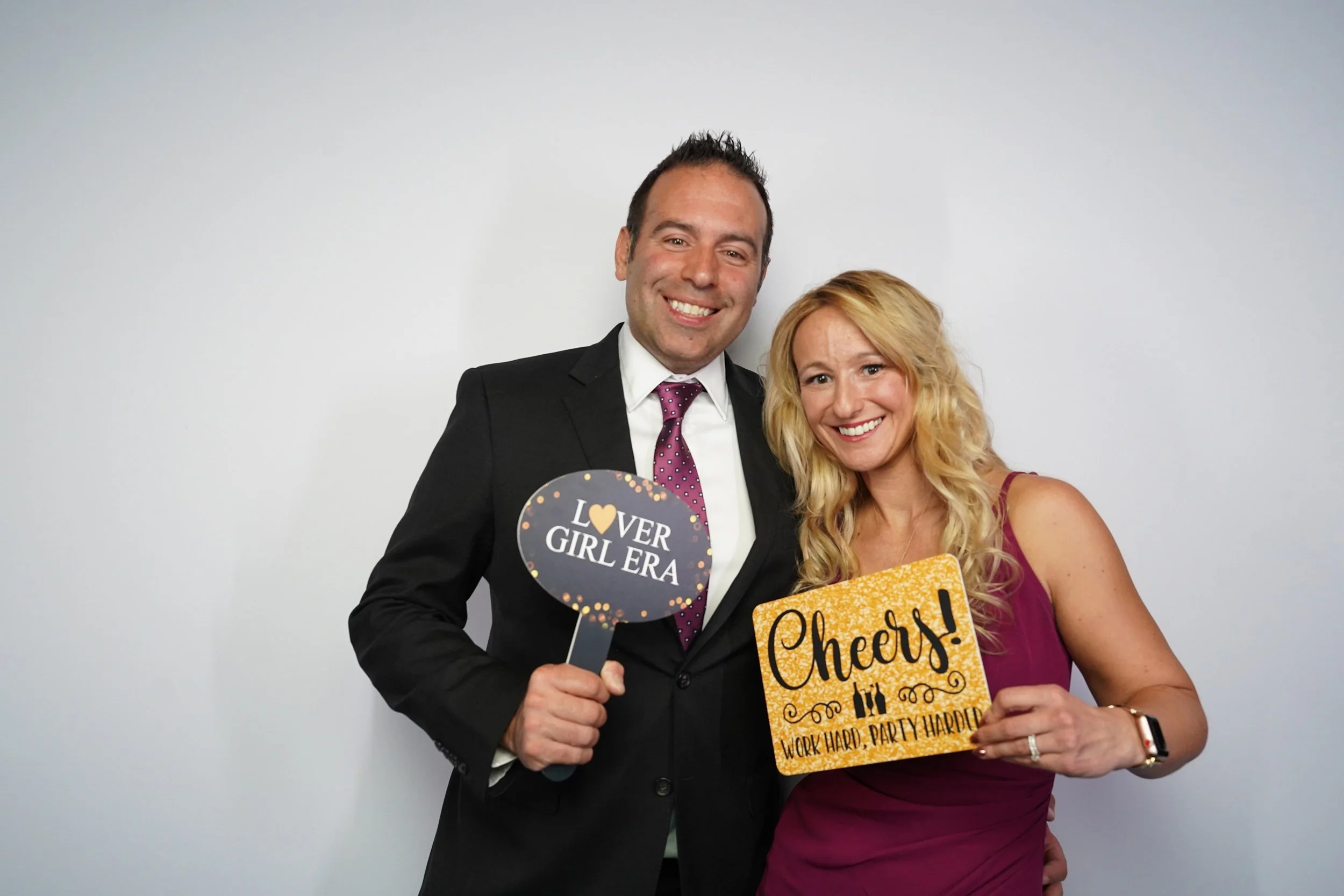 A smiling man in a suit and a woman in a sleeveless dress posing together, holding signs with celebratory messages. The man holds a sign that reads, 'LOVER GIRL ERA,' and the woman holds a sign that says, 'Cheers! WORK HARD, PARTY HARDER.' They are s