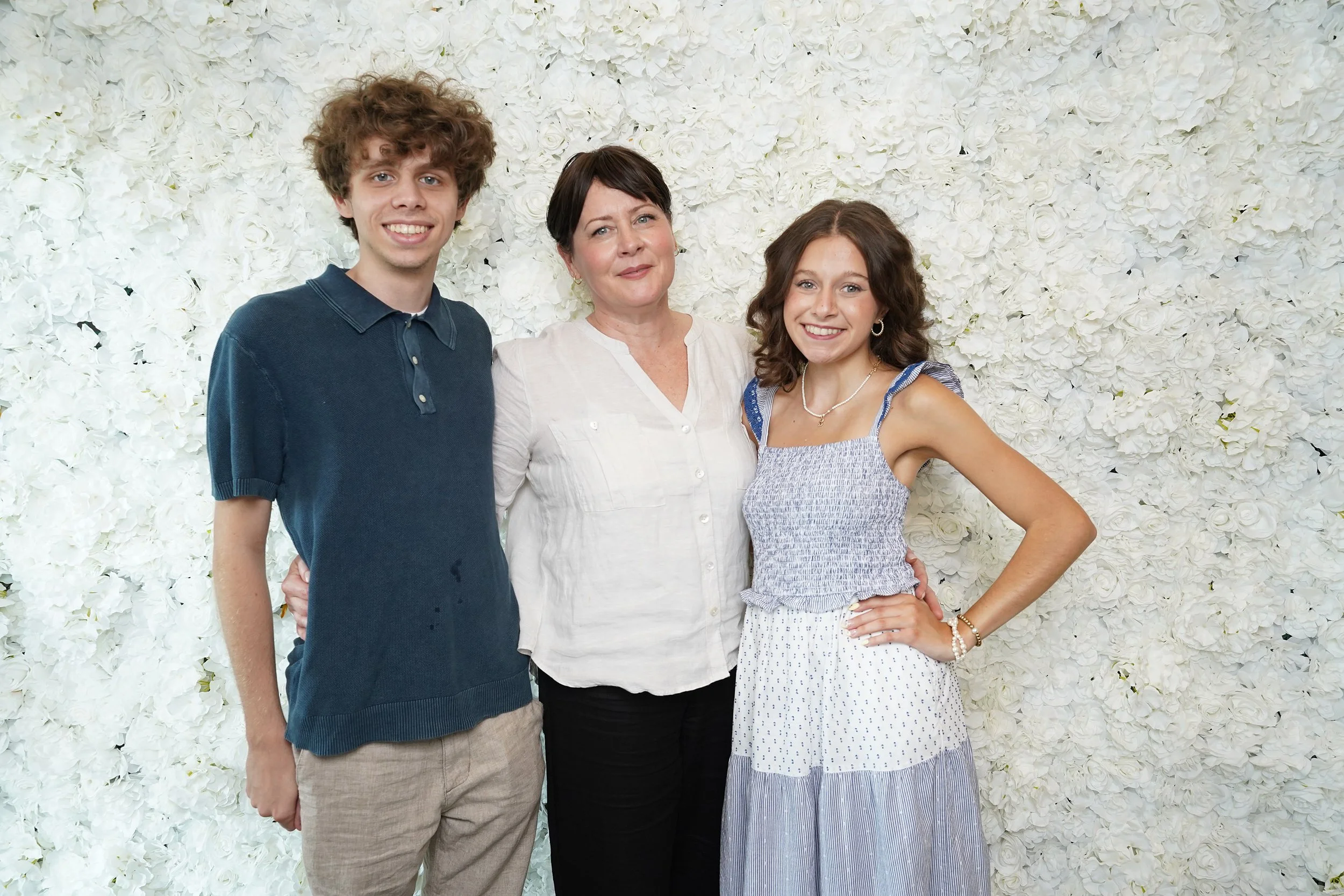 A woman standing with two young adults, all smiling in front of a white floral backdrop.