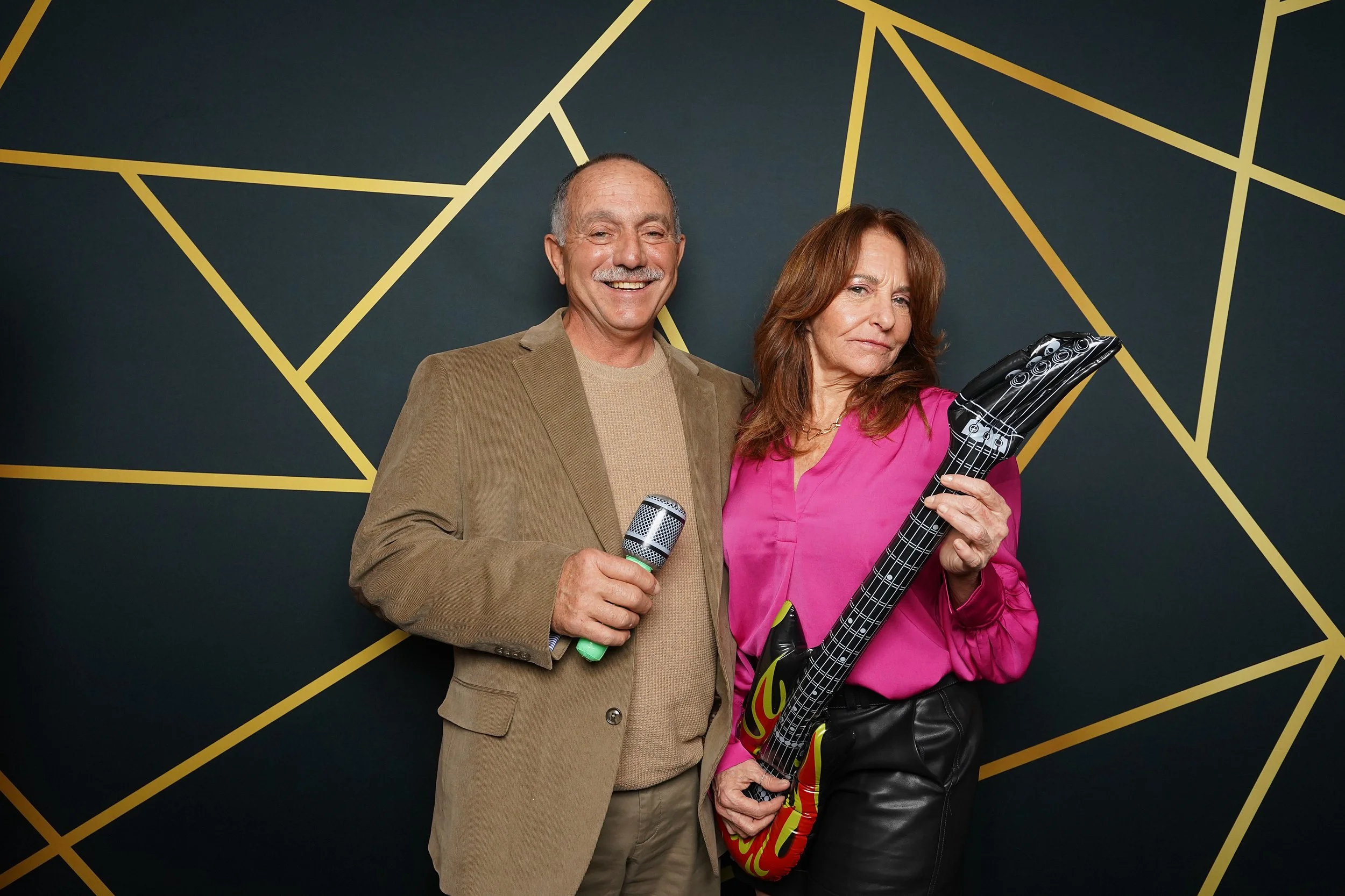 A smiling elderly man in a tan blazer holding a microphone, standing next to an older woman with long red hair in a pink blouse holding an inflatable guitar with flames design, against a dark wall with yellow geometric lines.