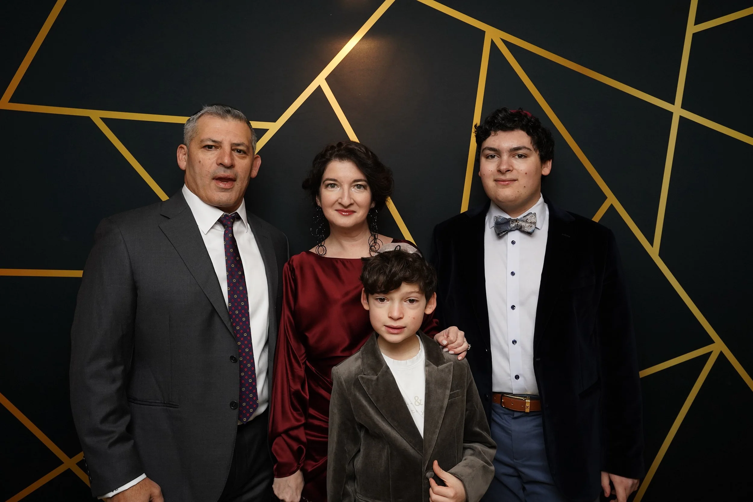 Family of four dressed in formal attire posing in front of a dark wall with gold geometric patterns.