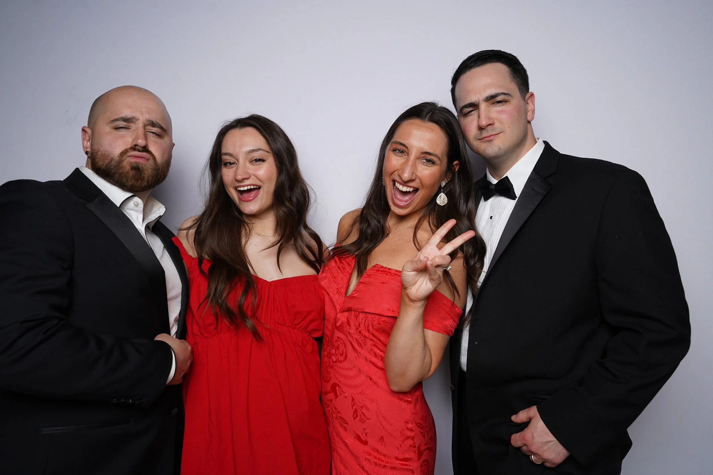 Group of four people dressed in formal attire, posing happily in front of a plain background, with a woman flashing a peace sign.