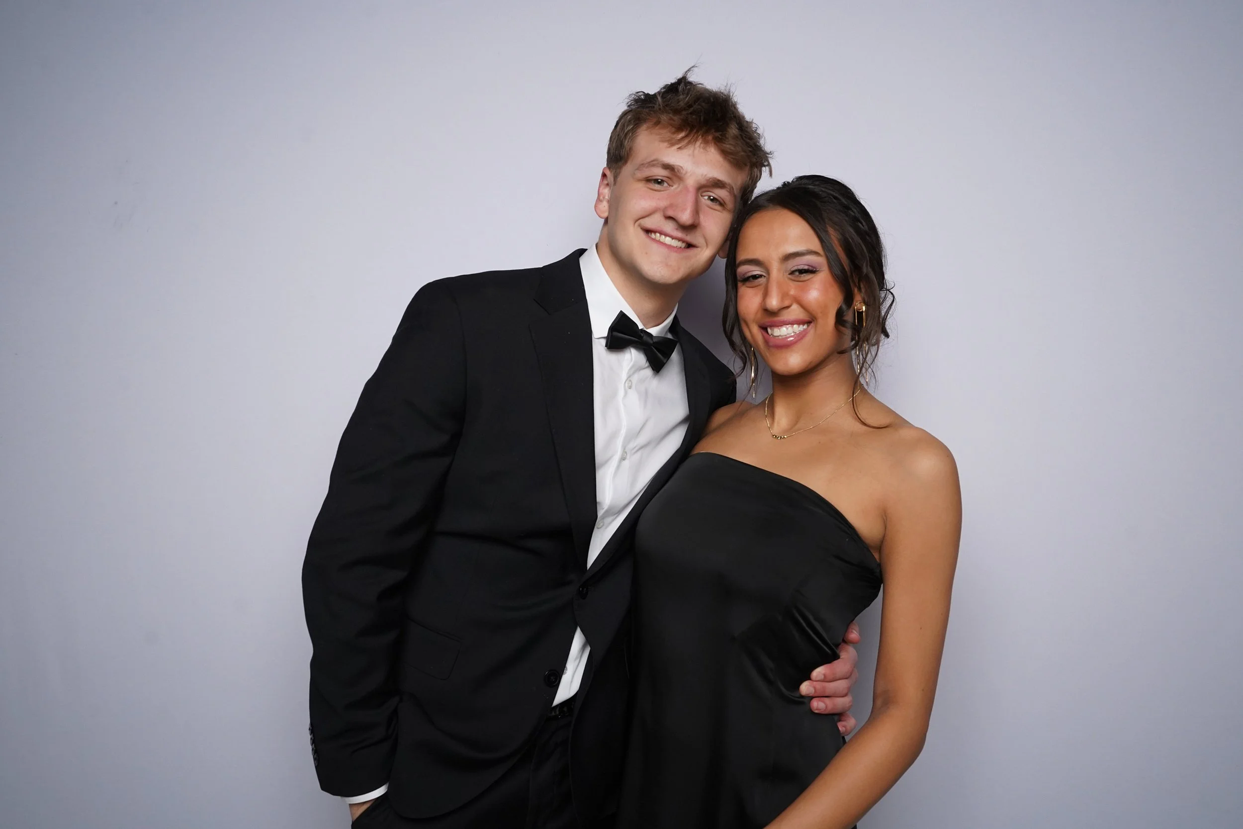A young couple dressed in formal attire smiling and posing together against a plain gray background.