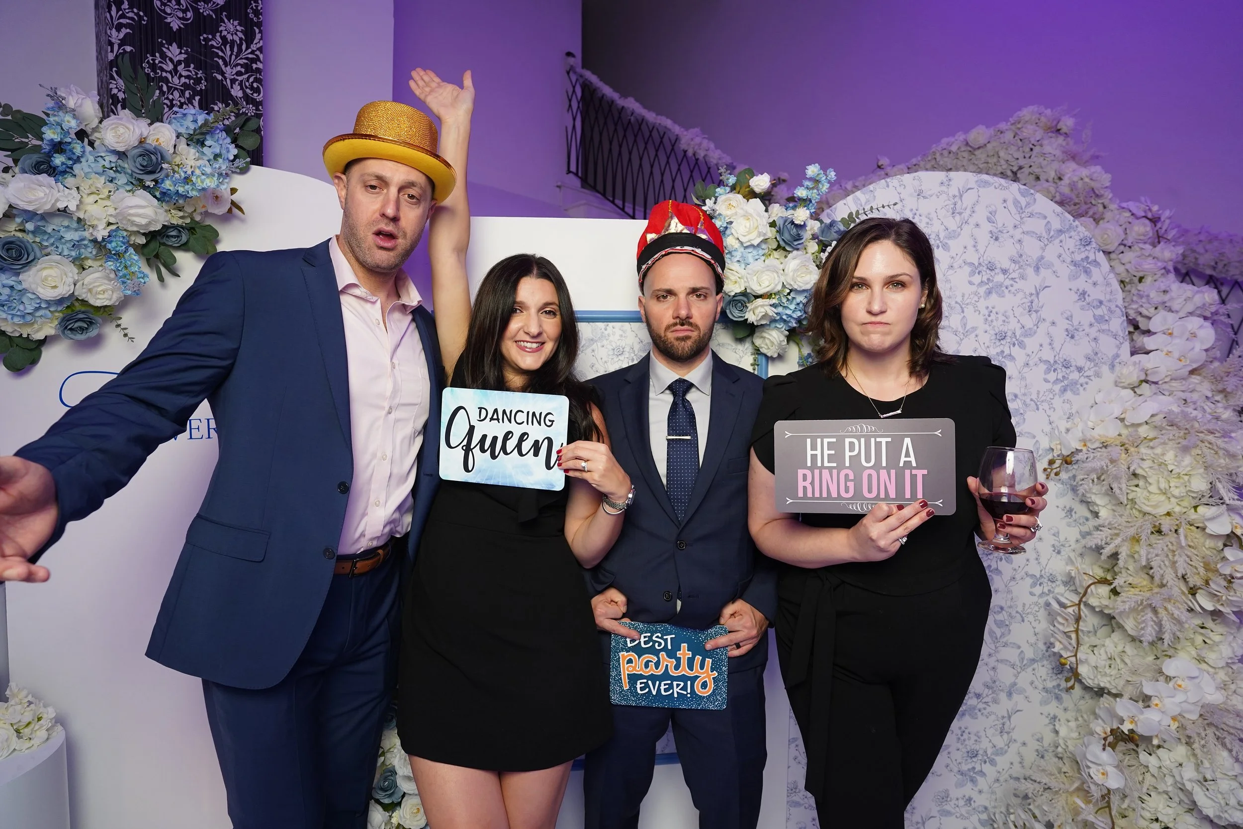 Four people standing at a wedding or party photo booth. They are dressed in formal attire and holding signs with humorous messages. One person is wearing a gold glittery hat, and another has a party hat. The background has floral decorations and purp