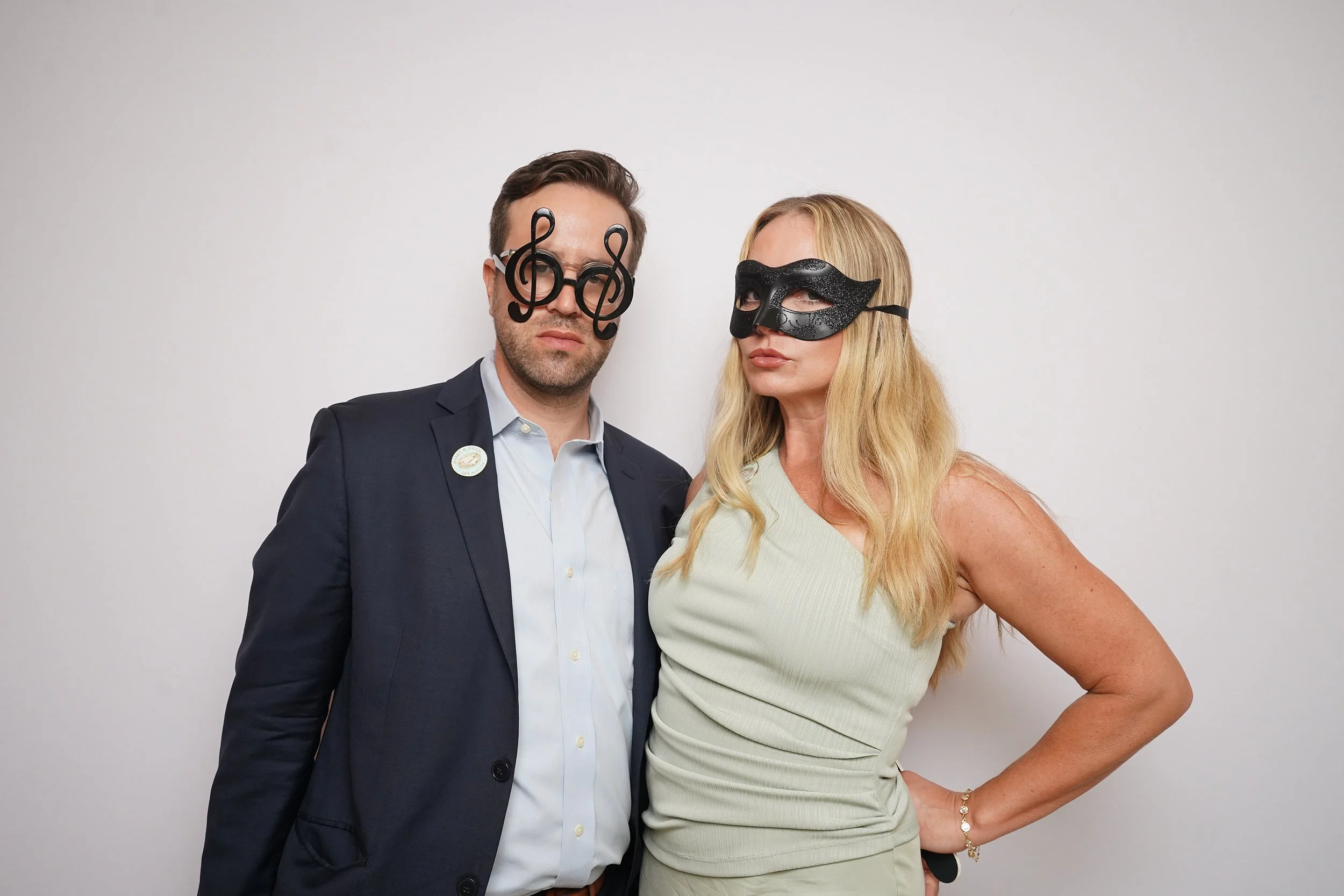 A man and a woman standing side by side against a plain white wall, both wearing masks; the man has novelty glasses shaped like musical notes and the woman has a black mask with a decorative pattern.