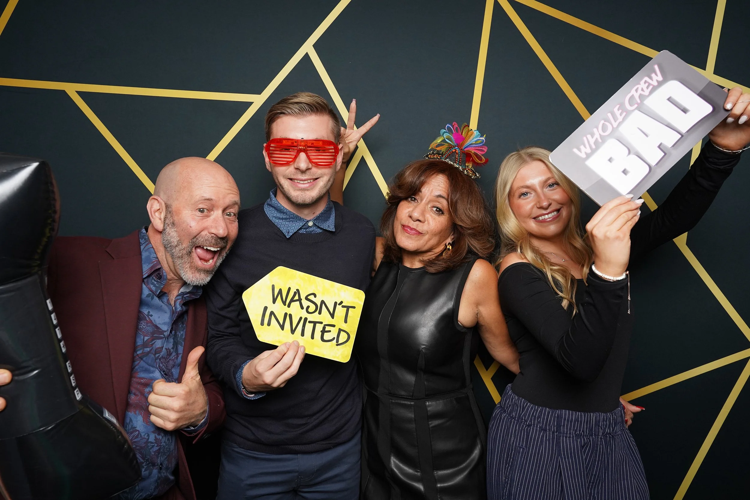 Group of people celebrating at a party with fun props, holding signs that say 'WASN'T INVITED' and 'NEW YEAR WHOLE CREW' in front of a geometric gold and dark background.