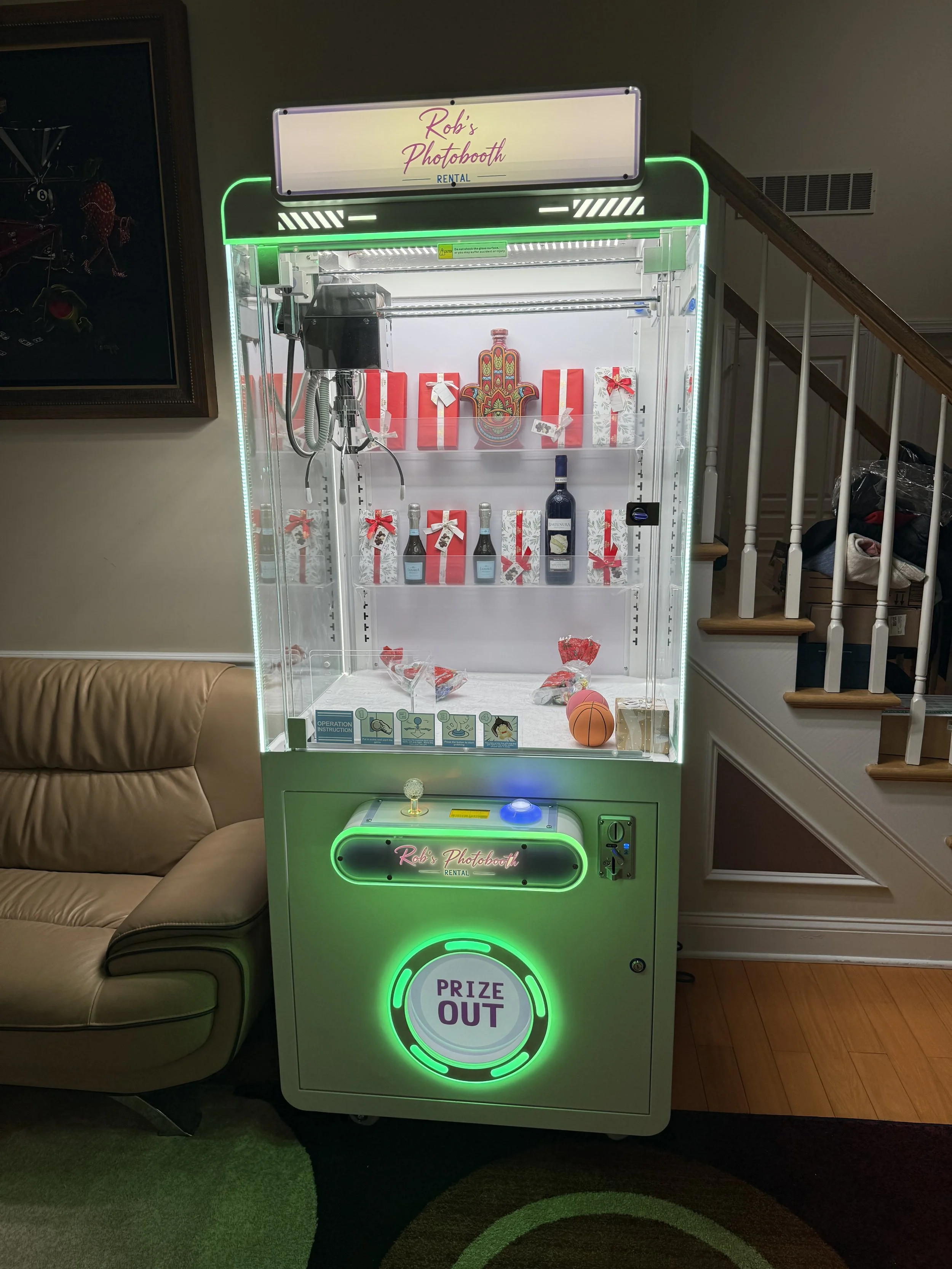 A claw machine labeled 'Rob's Photobooth' filled with wrapped gift boxes, a shell, a plastic basketball, and a small potted plant. The machine has bright green neon accents and a round green light labeled 'Prize Out' at the bottom. It is situated next to a beige couch and a staircase in a living room.