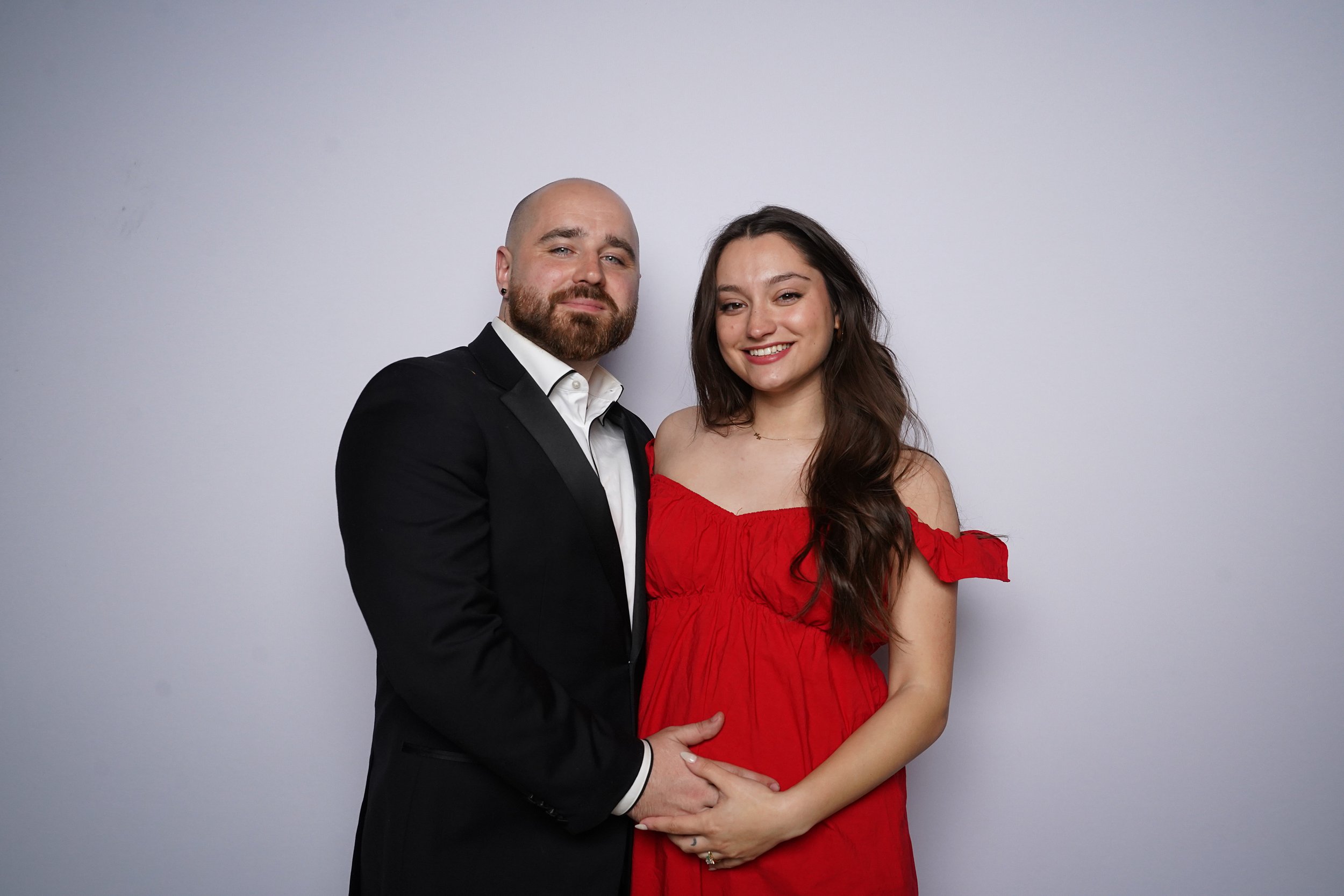 A couple posing together, the man in a black tuxedo and the woman in a red off-shoulder dress, standing against a plain light-colored background.