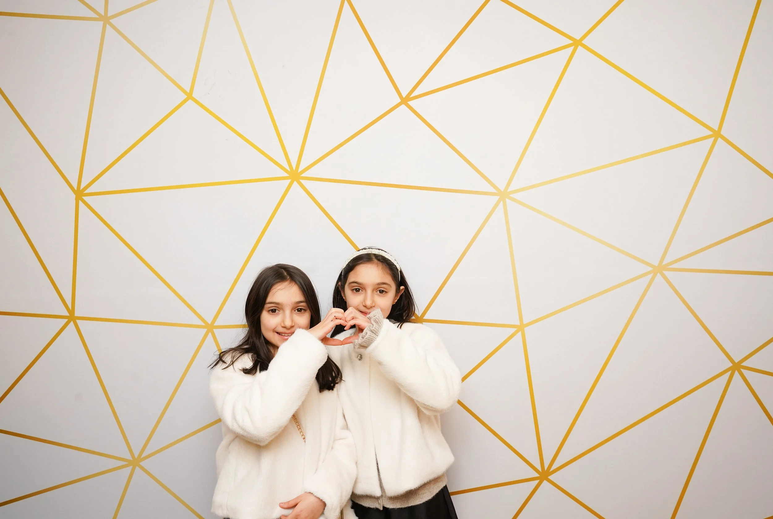 Two young girls in white furry coats, making a heart shape with their hands, standing against a white wall with a gold geometric pattern.