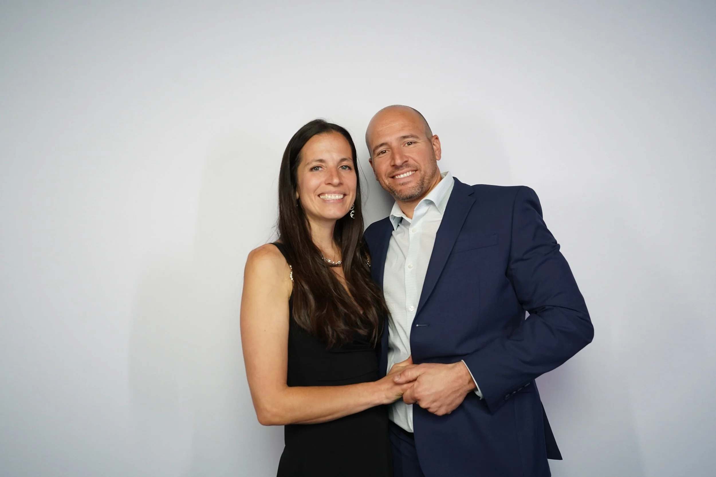A smiling woman in a black dress and a smiling man in a dark blue suit holding hands in front of a plain white background.
