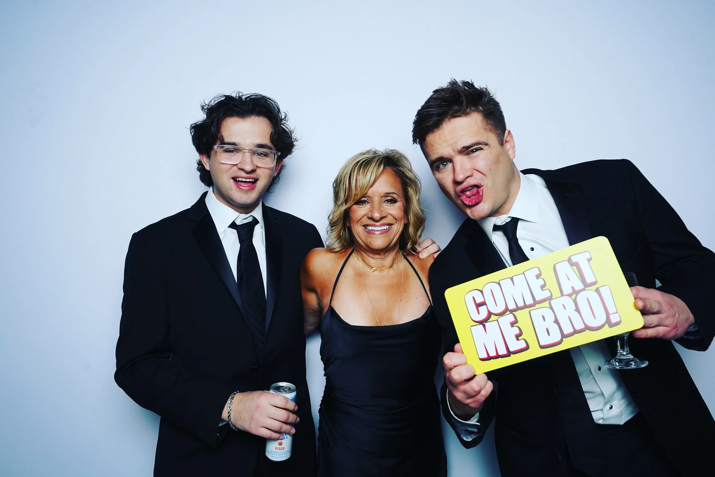 Three people dressed in formal attire at a party, with two men in suits and a woman in a black dress, standing against a plain white background. One man holds a drink and the other holds a yellow sign that says "COME AT ME BRO!"