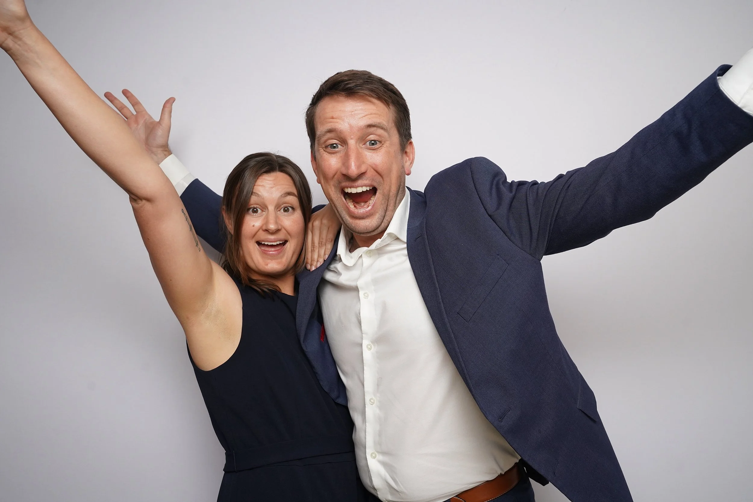 A man and a woman are celebrating happily, smiling broadly with arms raised, against a plain background.