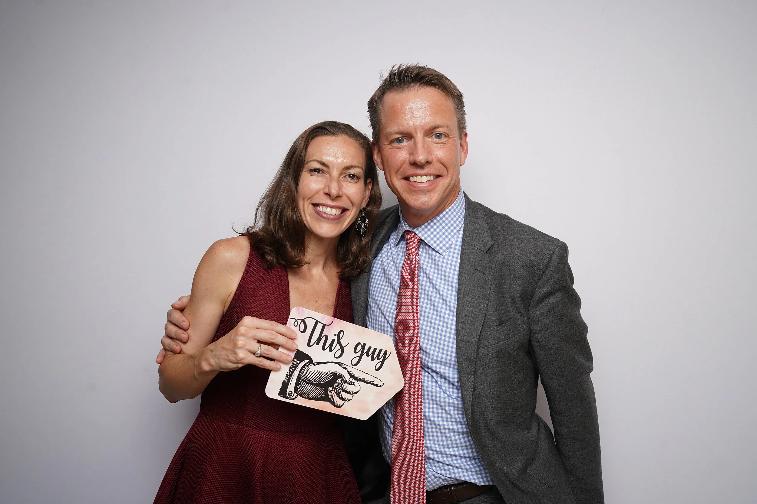 A woman and a man dressed in formal attire, smiling, with the woman holding a sign that says 'This guy' pointing to the man, against a plain white background.