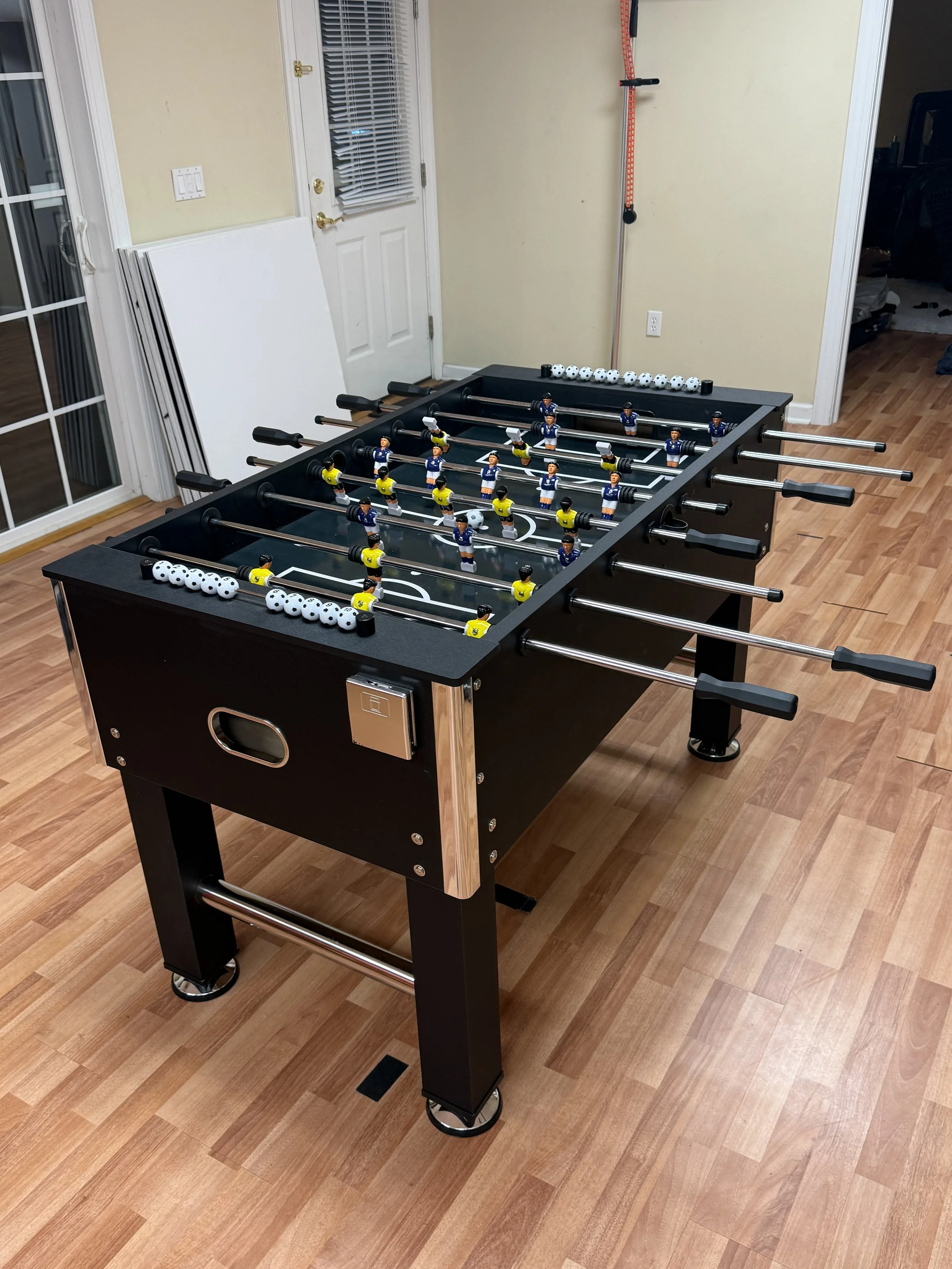 A foosball table in a room with wooden flooring, with white and black players arranged on rods and small white balls at each end of the table.
