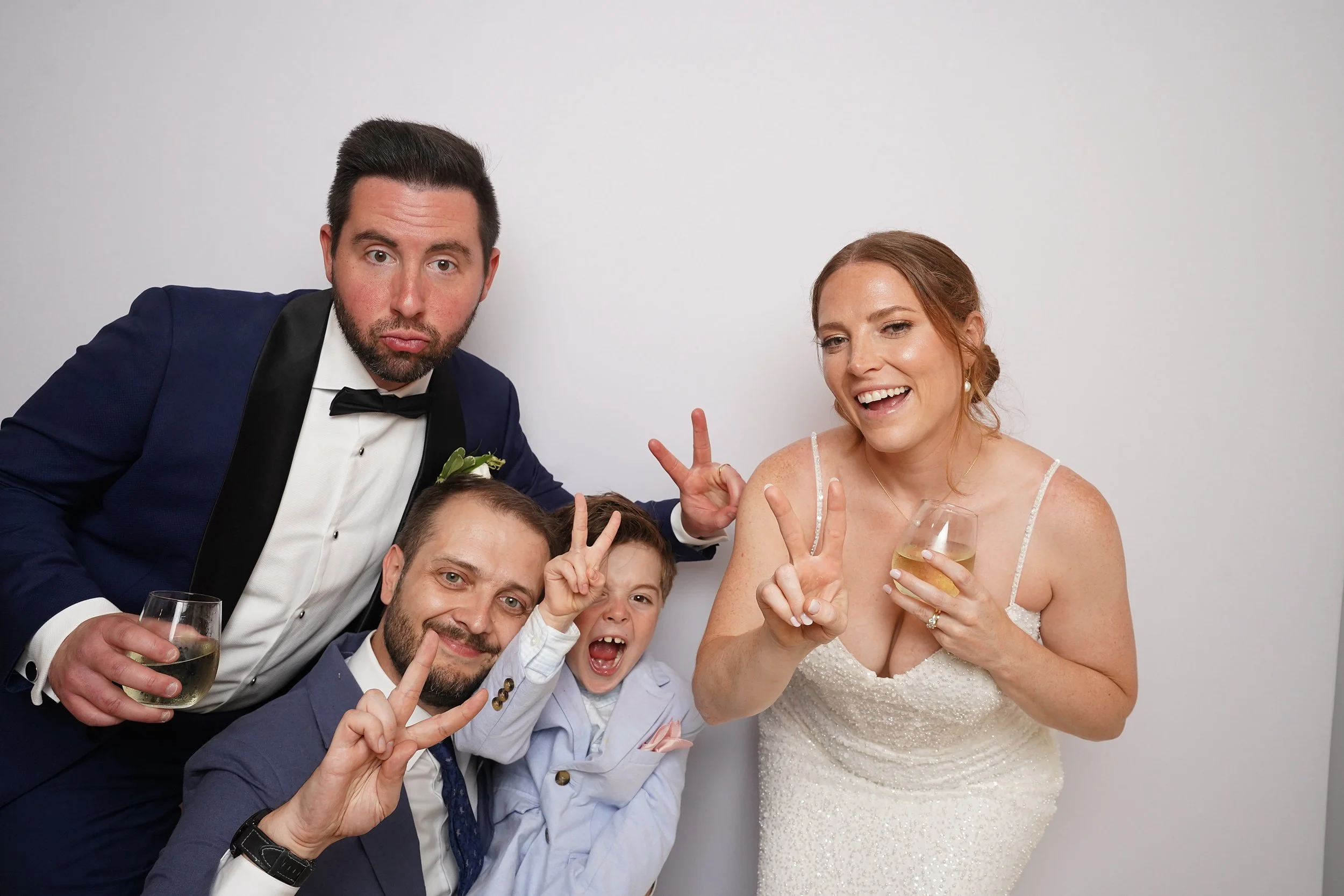 Group of four people celebrating at wedding, two men and a woman in a wedding dress, all making peace signs, holding drinks, against a plain white background.