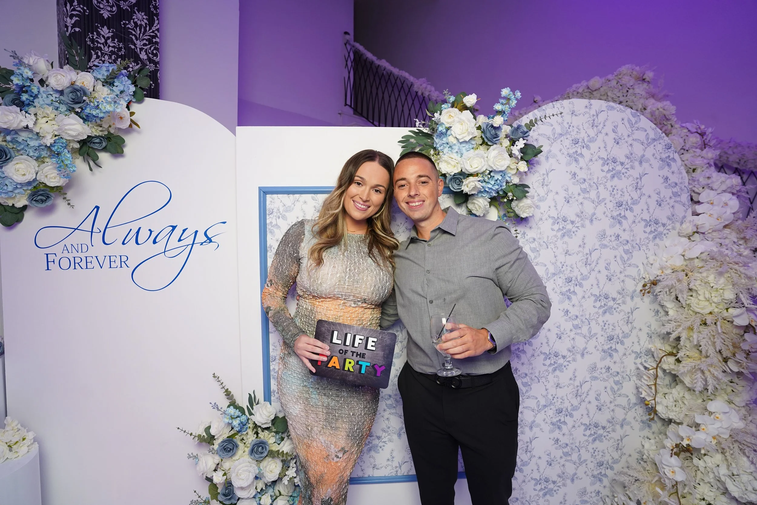 A smiling woman and man pose together at a decorated event backdrop, with floral arrangements and the words 'Always and Forever' written on it. The woman holds a sign that says 'Life of the Party', and the man holds a drink.