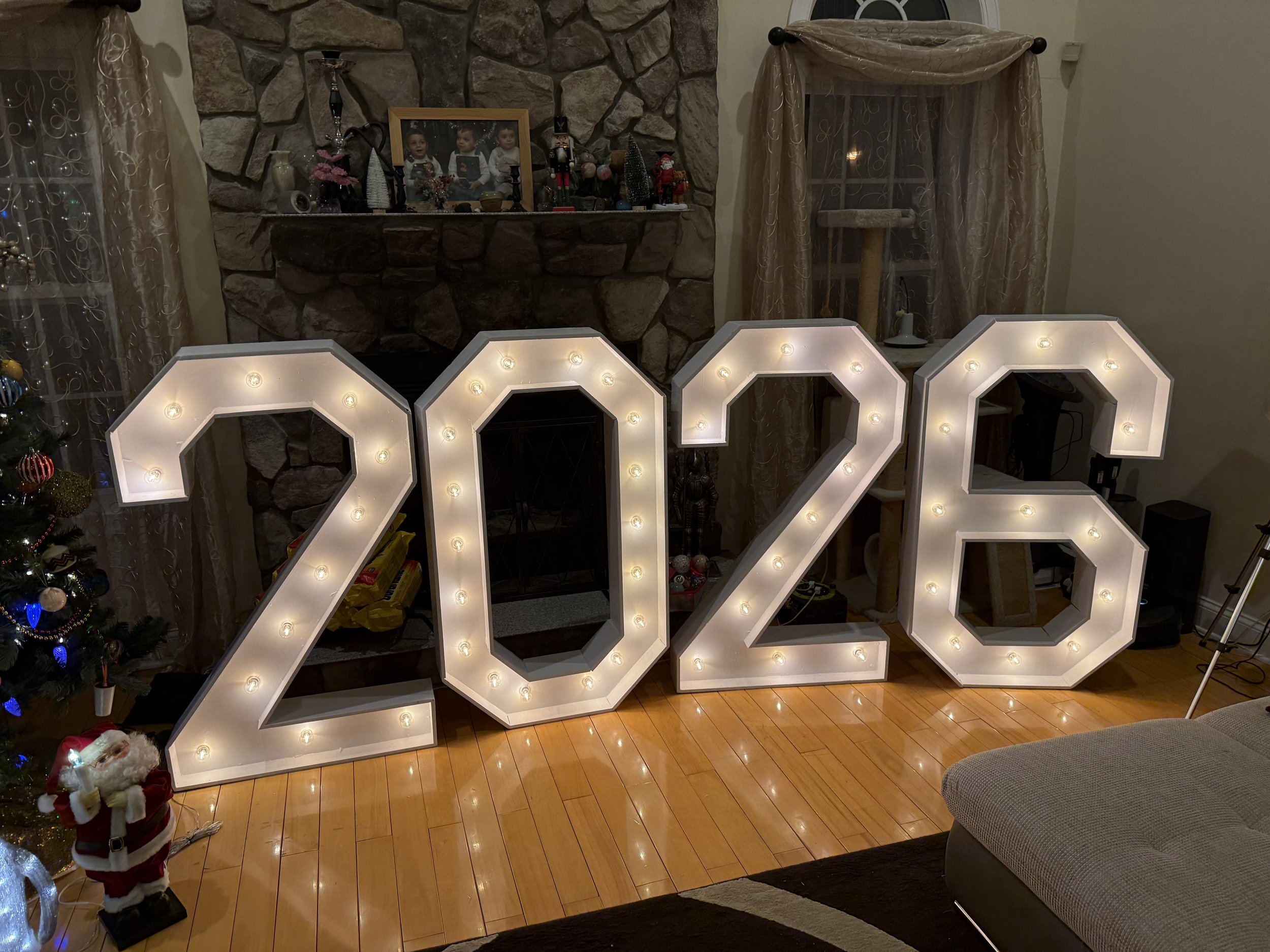 Large illuminated numbers display reading '2023' in a living room decorated for Christmas, with a Christmas tree on the left and a stone fireplace at the center.